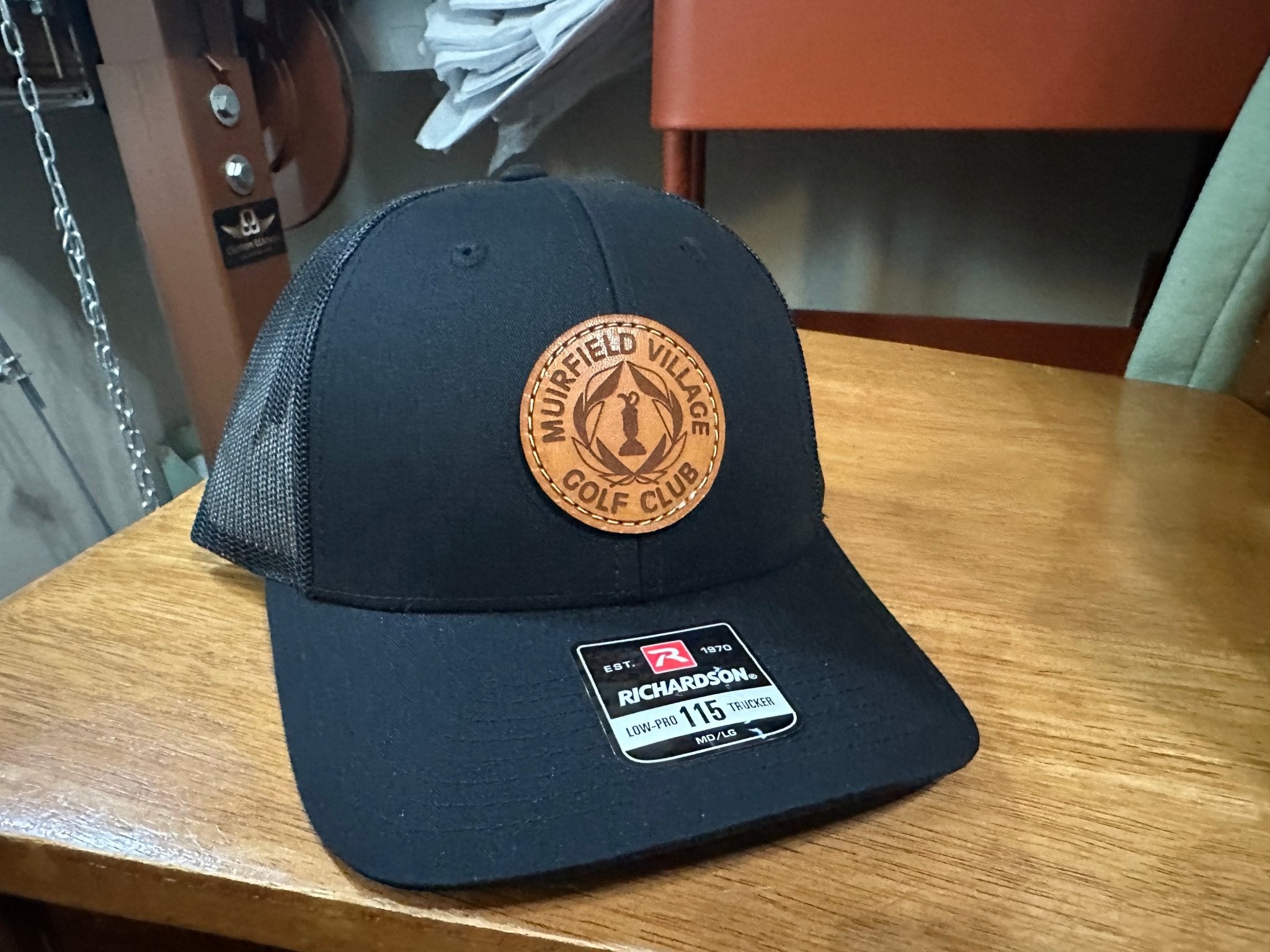 Black Richardson 115 hat with a round custom leather patch sewn on, featuring the text ‘Muirfield Village Golf Club’ with a gold trophy and wreath design in the center. A stylish and high-quality hat perfect for golf enthusiasts and fans of the club