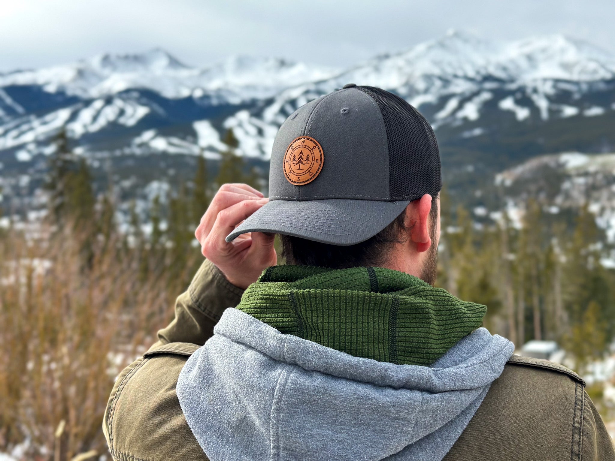 Man wearing the Lost Pine hat backwards, looking out over the snowy mountains of Breckenridge Ski Resort. The hat features a circular leather patch with a compass design and pine trees in the middle, symbolizing adventure and outdoor exploration