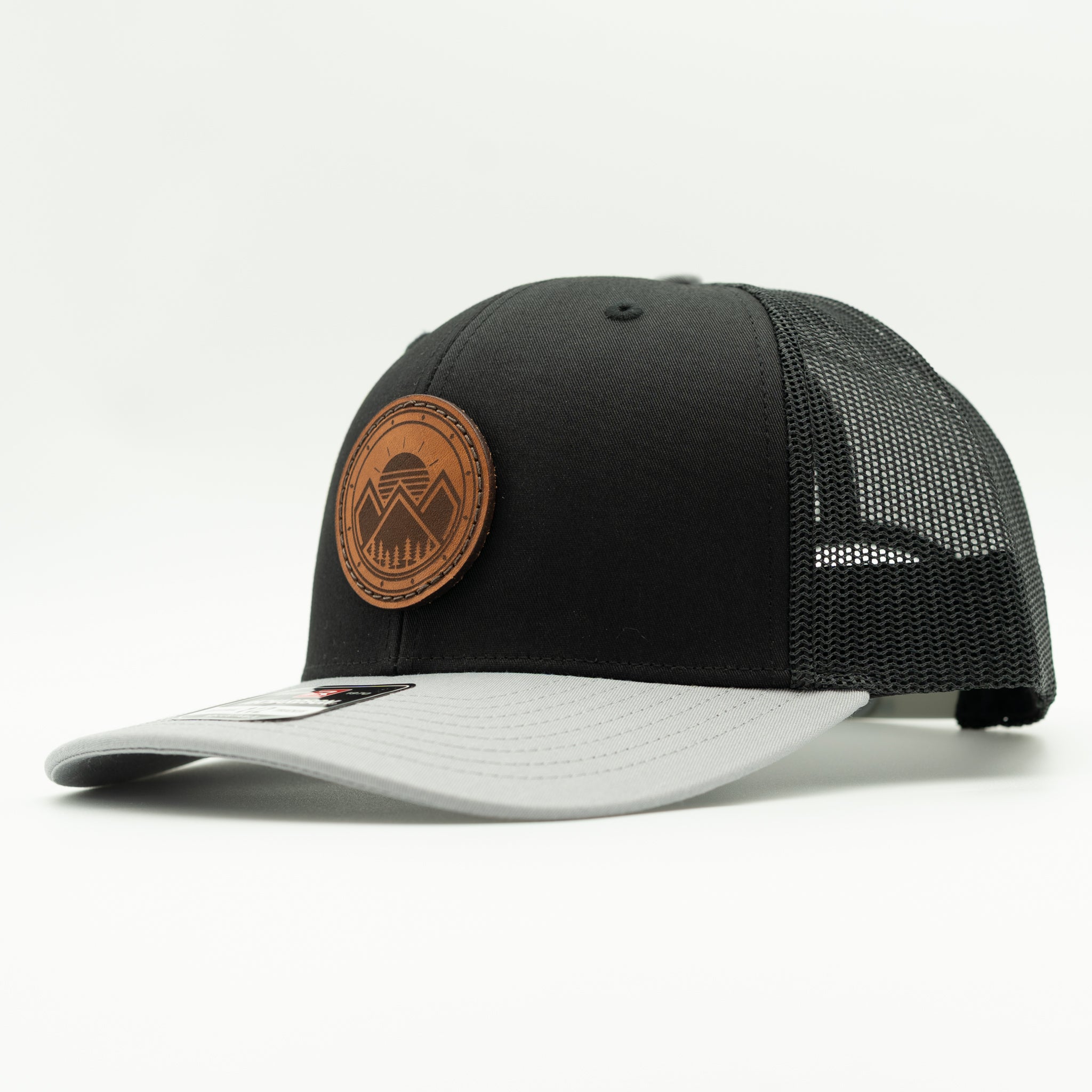 A black trucker-style cap with a light gray visor and black mesh back, featuring a circular brown leather patch on the front with a mountain logo.
Color: Black/Grey Combo