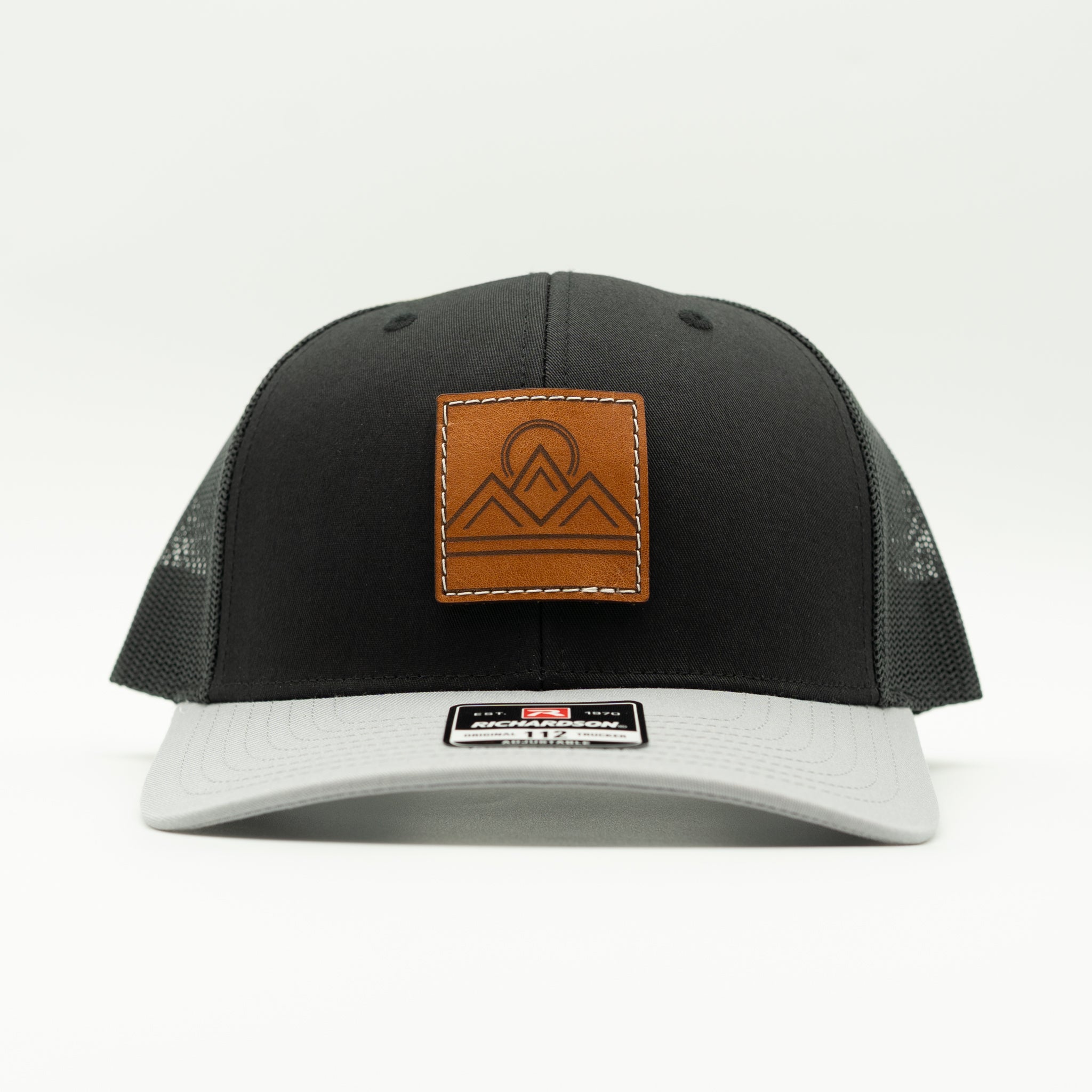 Front view of Lunar Ridge black and grey combo Richardson 112 trucker hat with square leather mountain patch