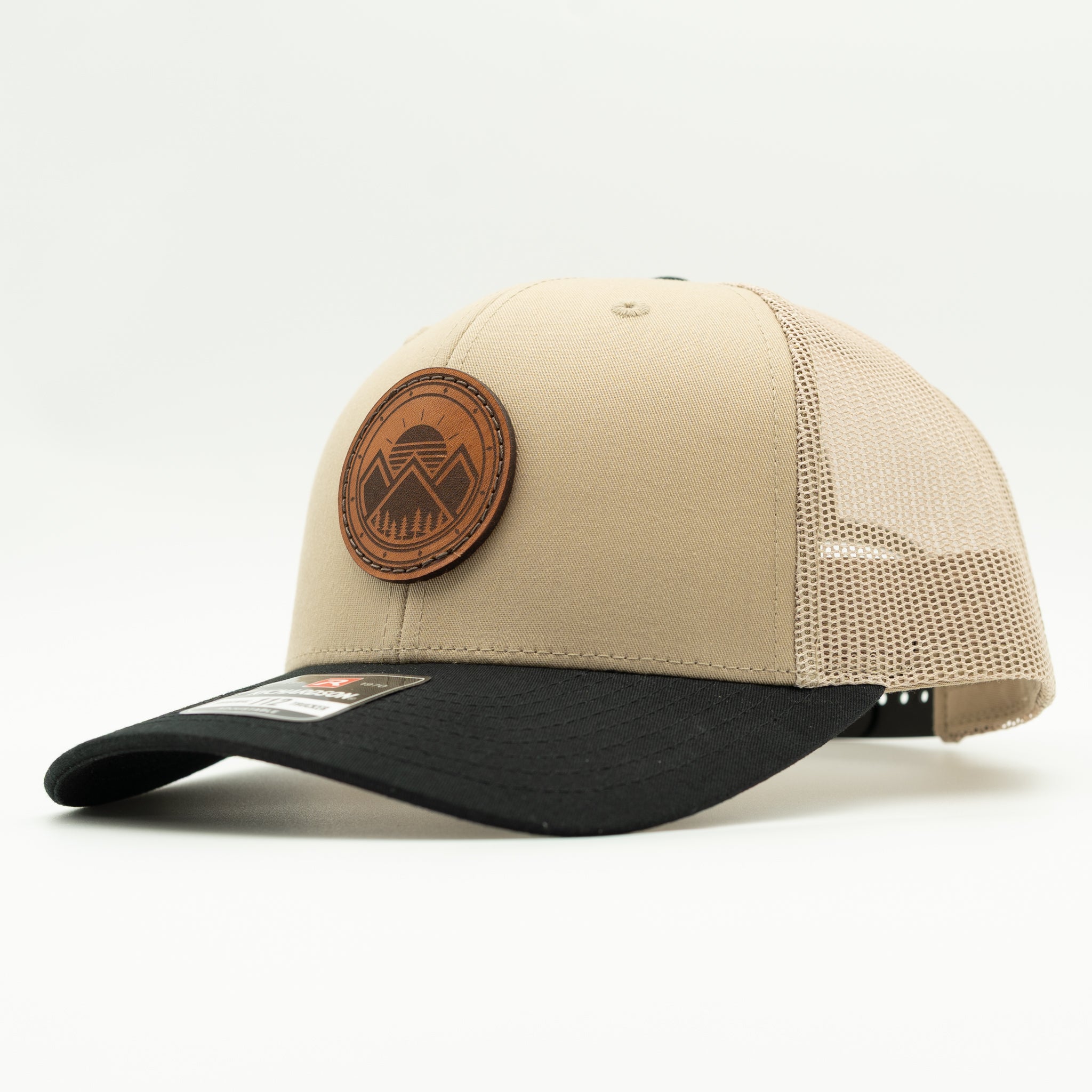 A trucker-style baseball cap with a beige front panel and black brim, featuring a circular brown leather patch on the front. The back of the cap has a beige mesh back.
Color: Khaki/Black Combo