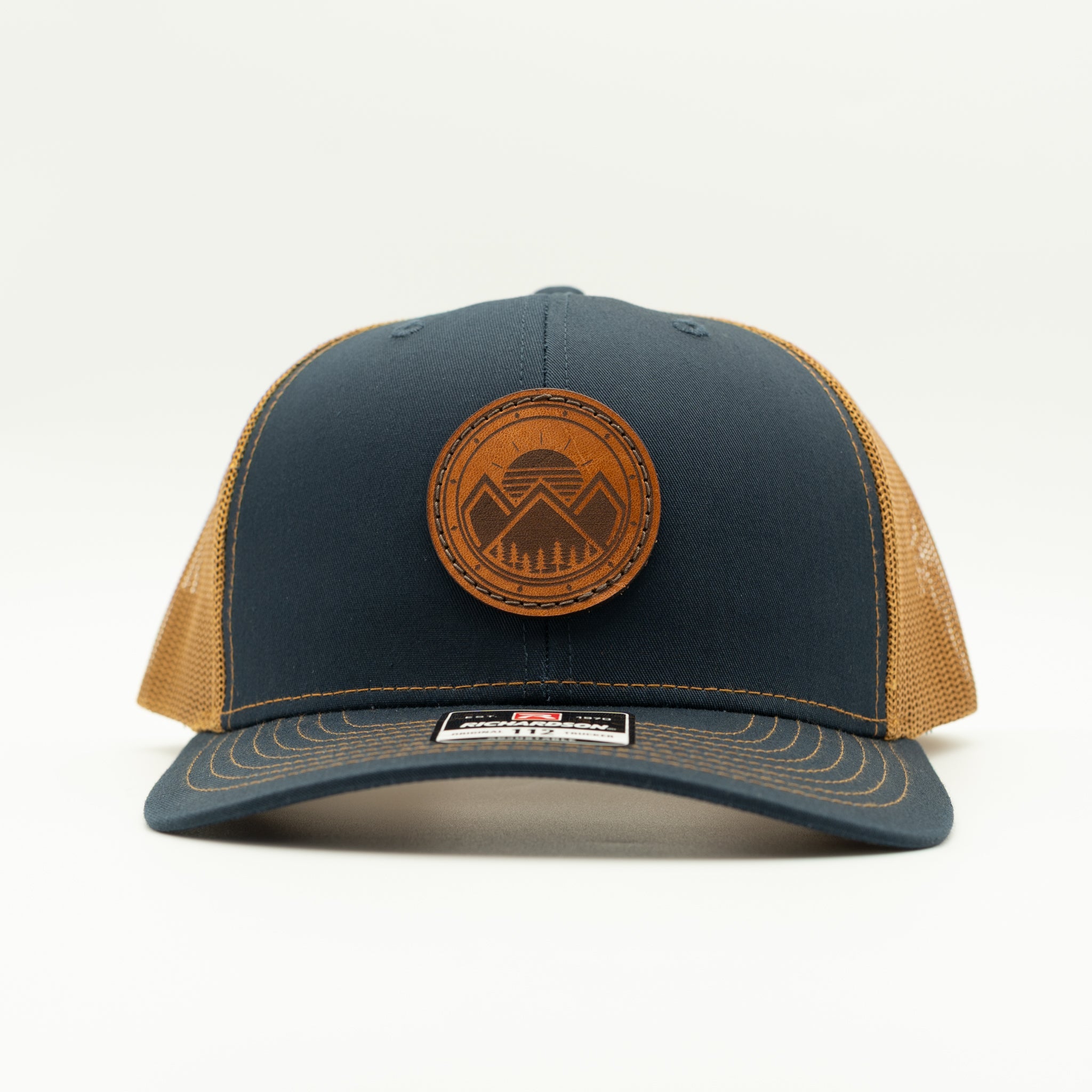 A navy blue trucker hat with tan mesh back panels and a circular brown leather patch on the front featuring a mountain and sun design.
Color: Navy/Caramel
