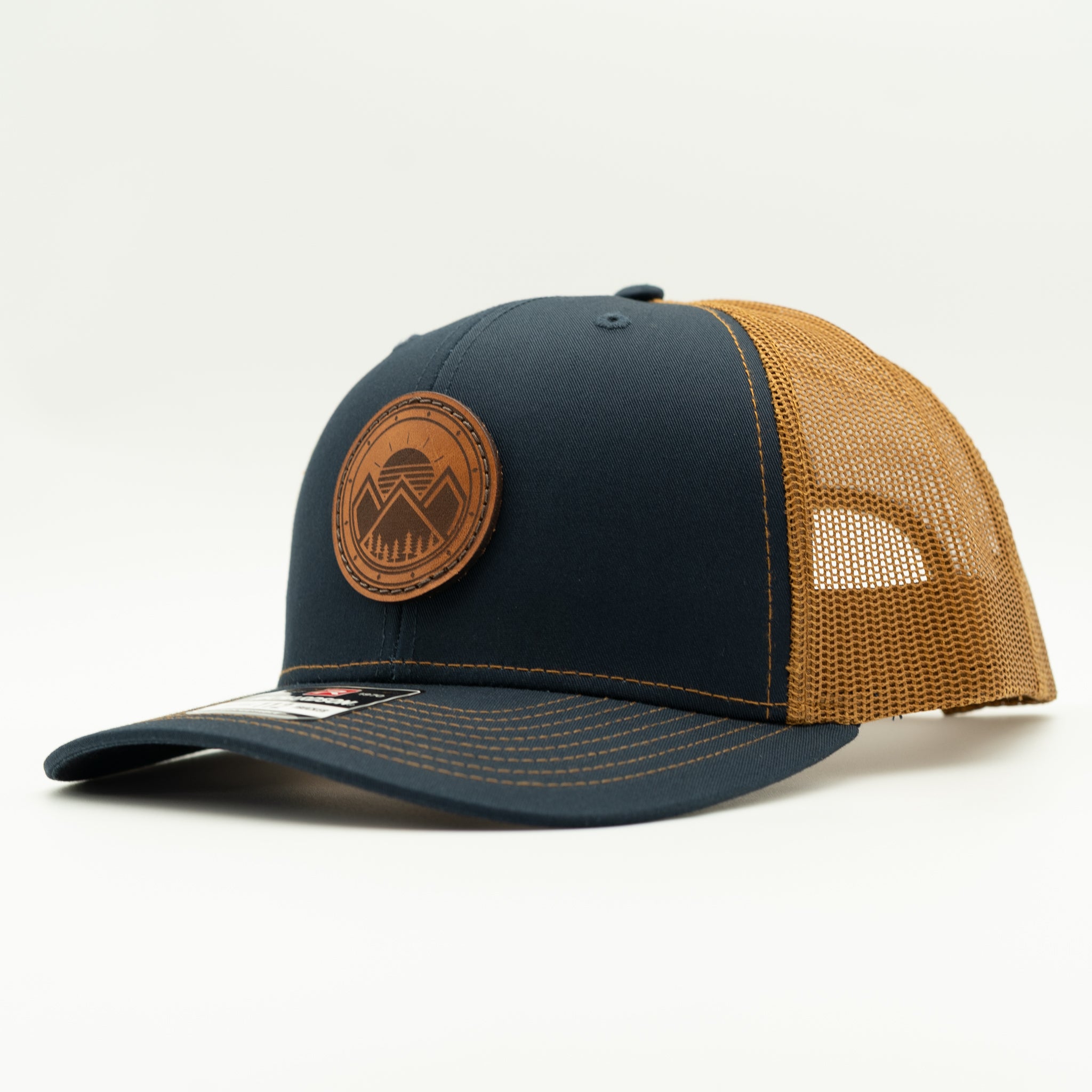 A structured trucker-style baseball cap with a dark navy blue front panel and brim, tan mesh back panels, and a circular brown leather patch on the front featuring a mountain design.
Color: Navy/Caramel
