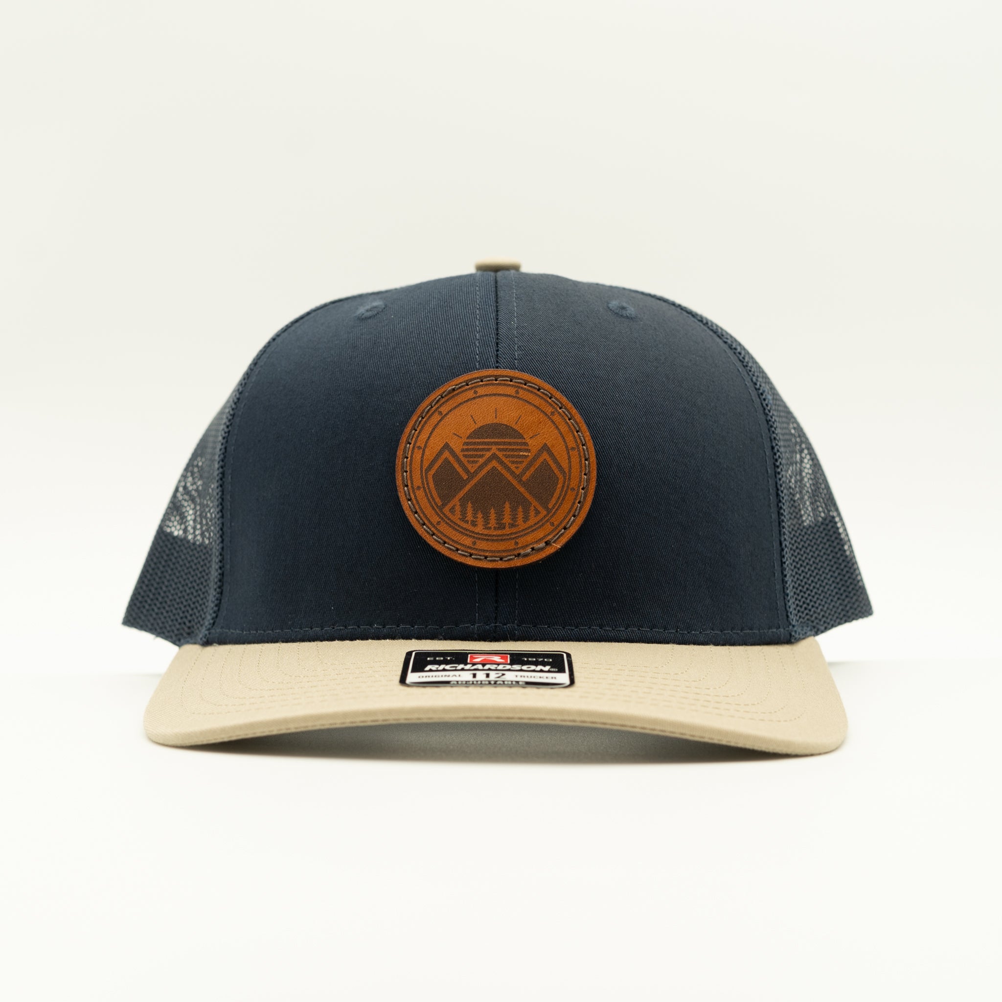 A navy blue trucker cap with a beige flat brim, featuring a circular brown leather patch on the front with a mountain design. The back panels are made of navy mesh, and a black size label is visible on the brim.
Color: Navy/Khaki Combo