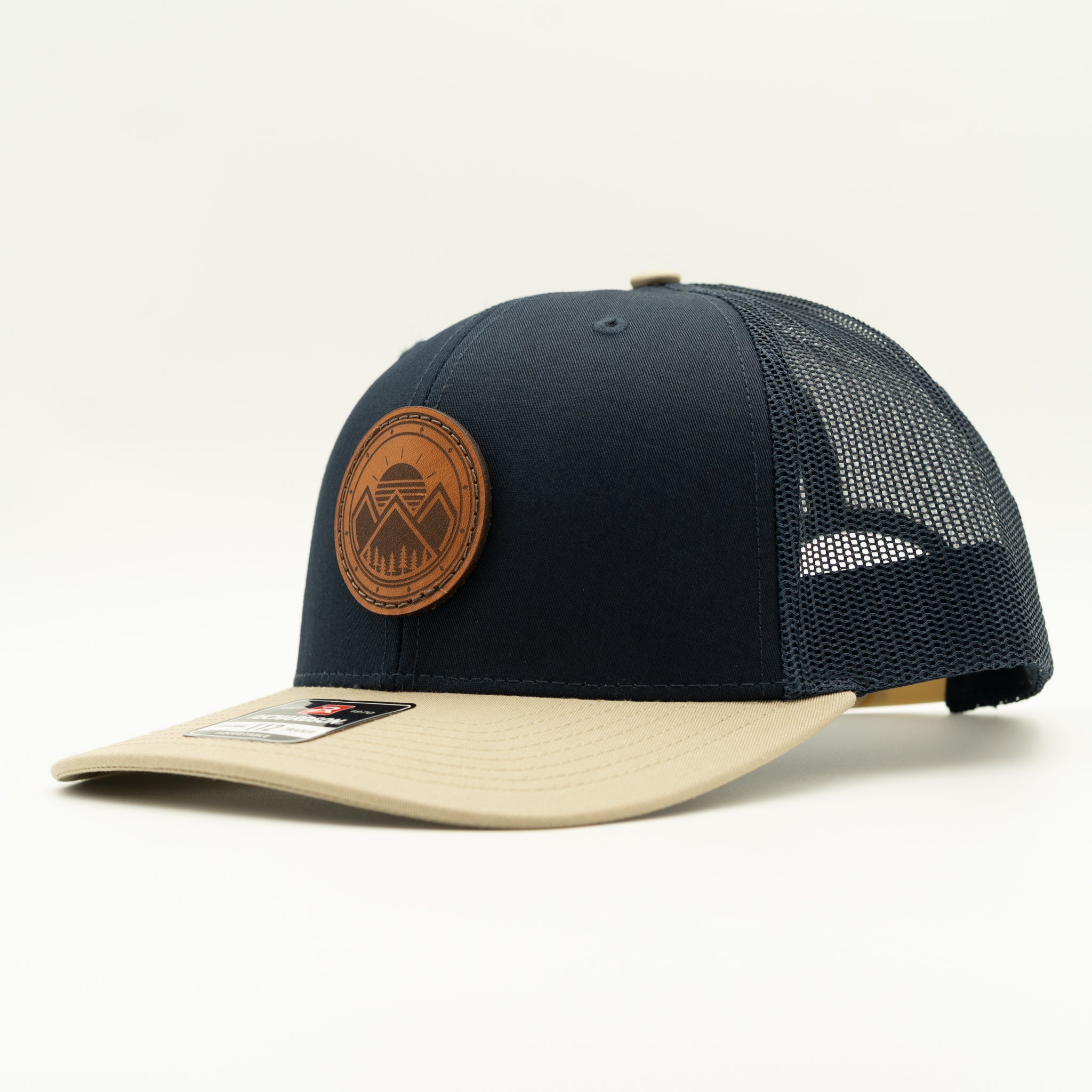A structured trucker cap with a dark navy blue front panel and mesh back, a beige flat brim, and a circular brown leather patch on the front.
Color: Navy/Khaki Combo