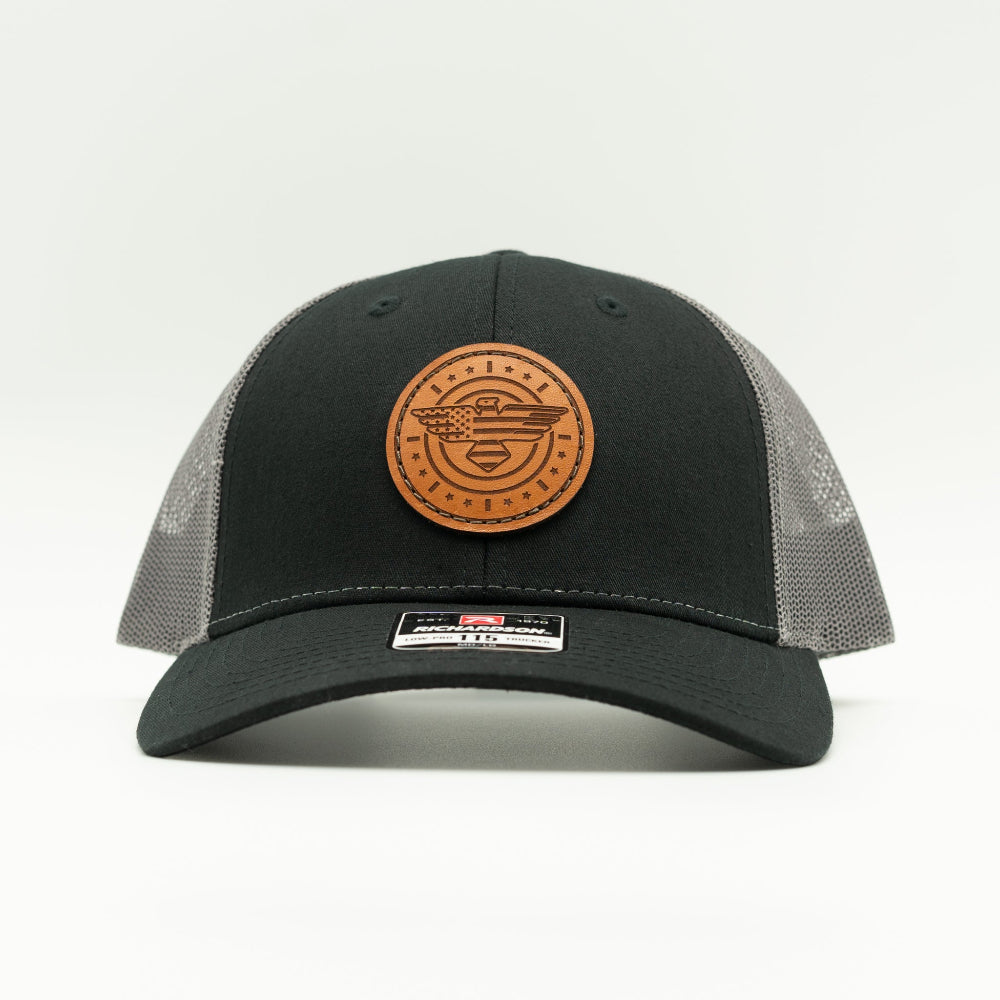 Front view of black and charcoal Richardson 115 low profile trucker hat with round American Eagle and flag leather patch, curved visor, and charcoal mesh back.
Color: Black/Charcoal