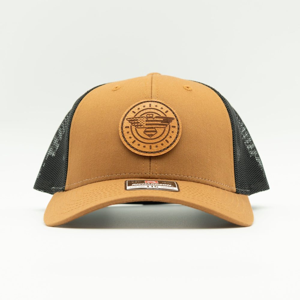 Front view of caramel and black Richardson 115 low profile trucker hat with detailed American Eagle and flag leather patch, curved visor, and black mesh back.
Color: Caramel/Black