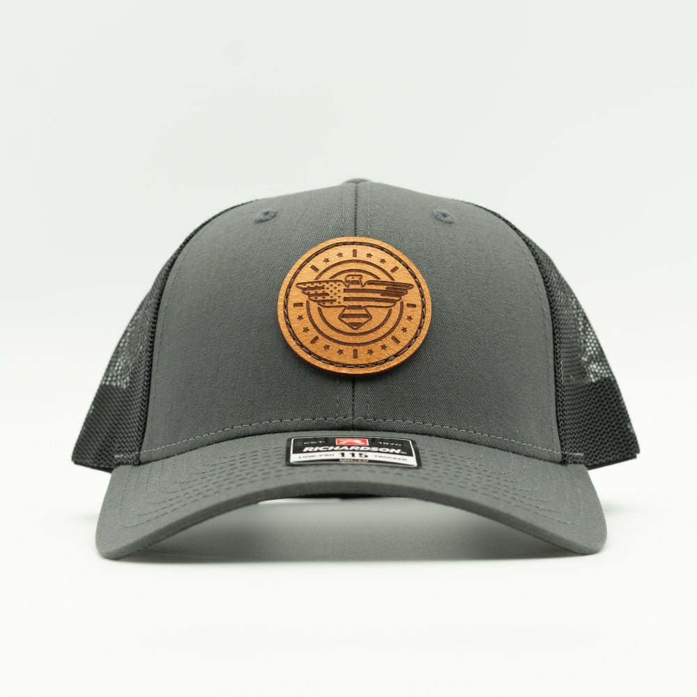 Front view of charcoal and black Richardson 115 low profile trucker hat with round American Eagle and flag leather patch, curved visor, and black mesh back.
Color: Charcoal/Black