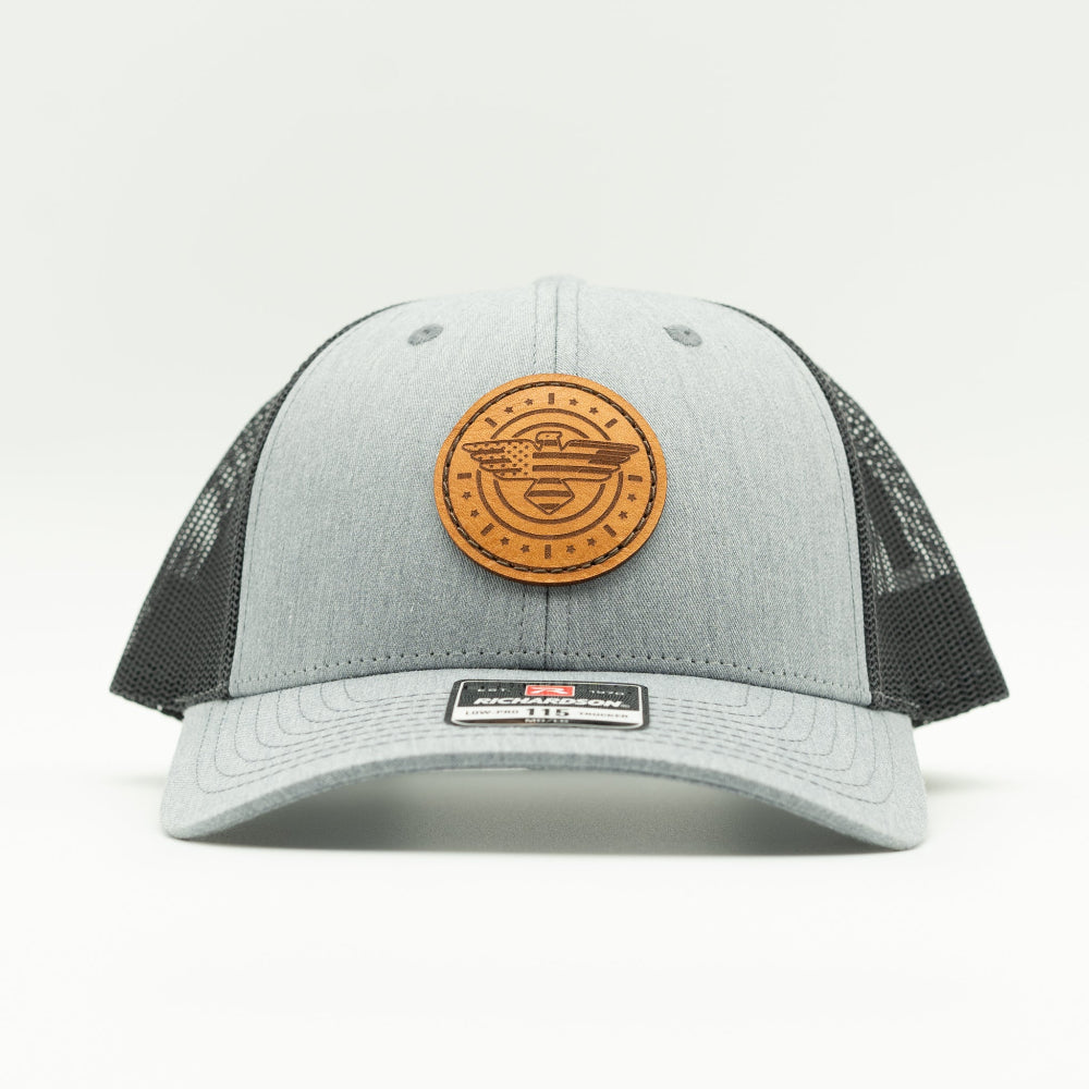 Front view of heather grey and charcoal Richardson 115 low profile trucker hat with engraved American Eagle and flag leather patch, curved visor, and dark mesh back.
Color: Heather Grey/Charcoal