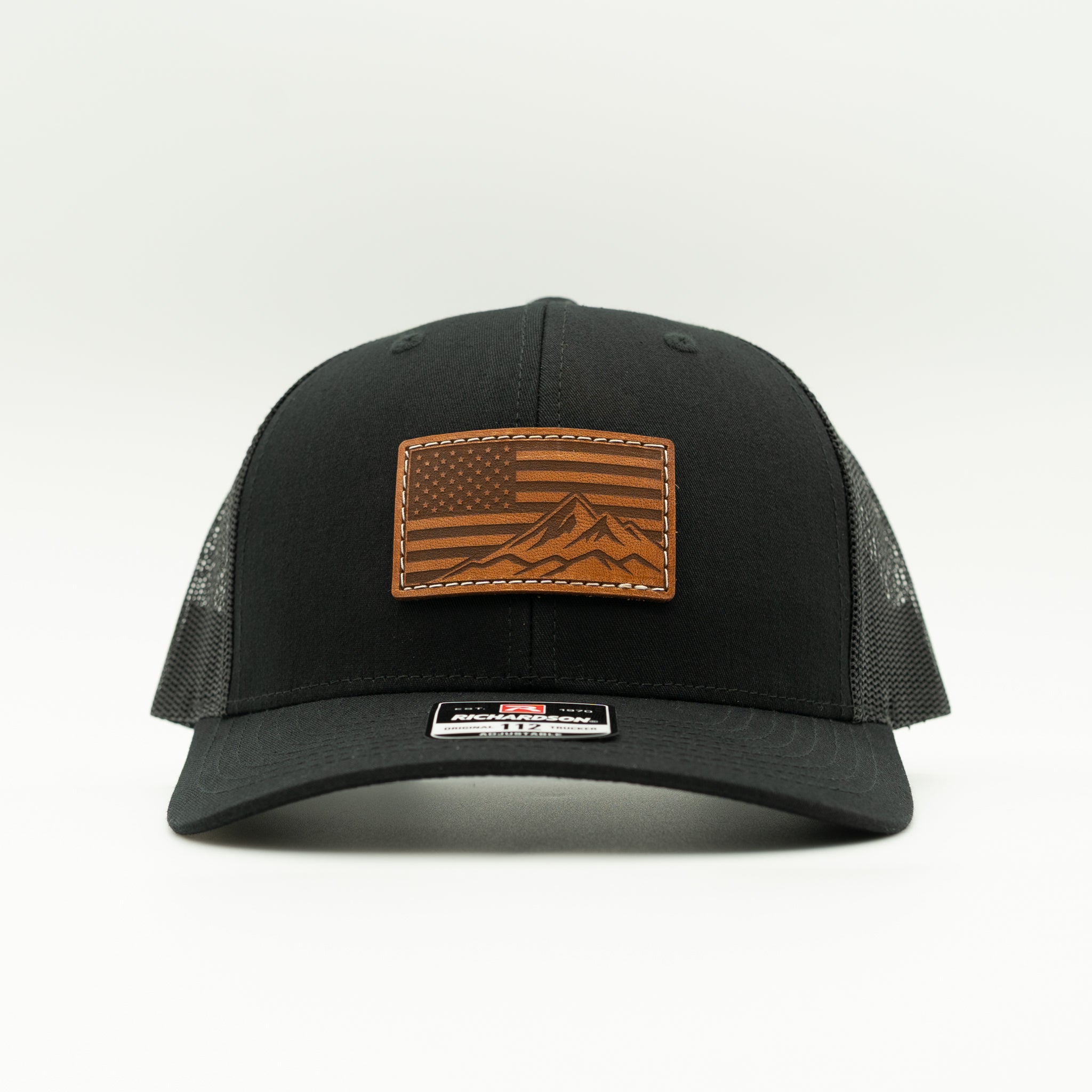 Front angled view of American Range leather patch trucker hat on solid black Richardson 112, with light brown American flag patch, curved bill, white stitching, mesh back, and snapback, a clean patriotic mountain hat for daily wear and travel.