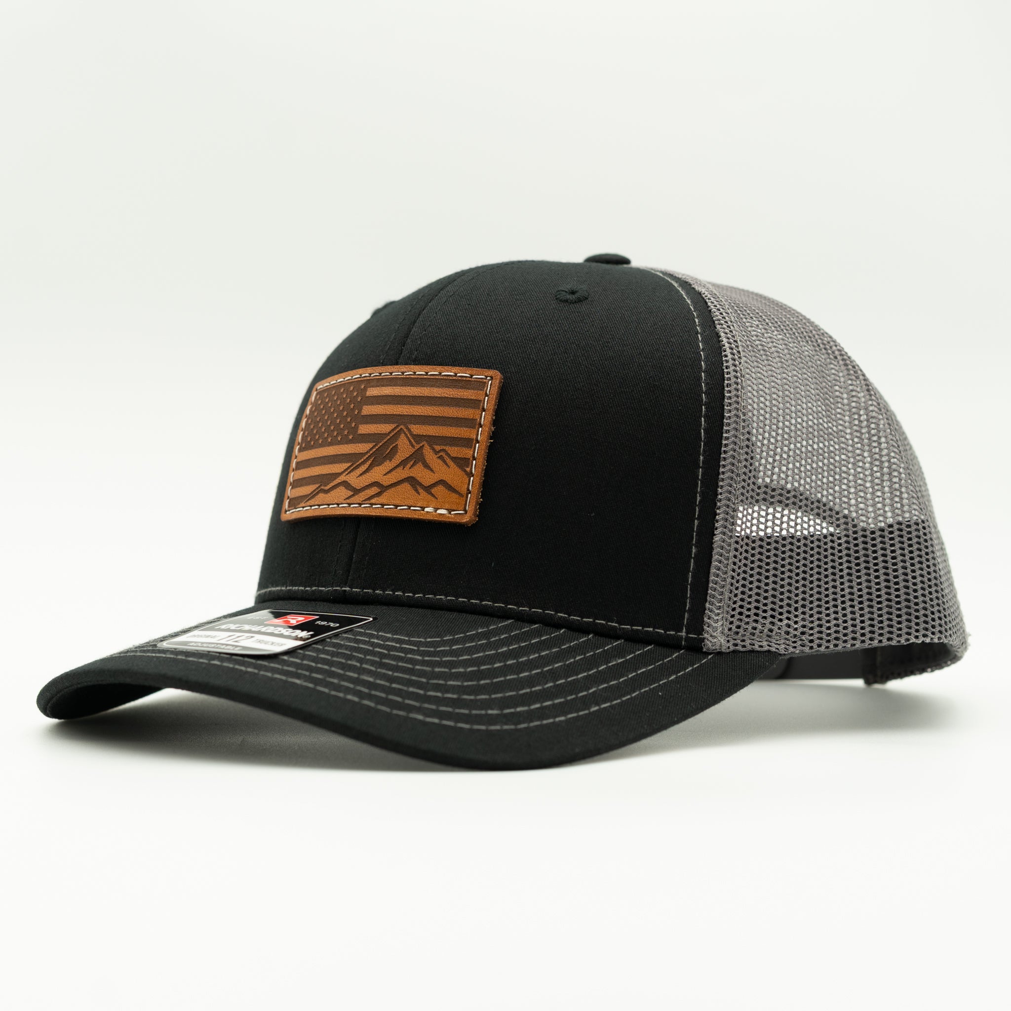 Side profile of American Range American flag leather patch hat on black and charcoal Richardson 112 trucker, showing patch, curved bill, mid profile fit, mesh back, and snapback, a versatile patriotic mountain hat for daily wear.