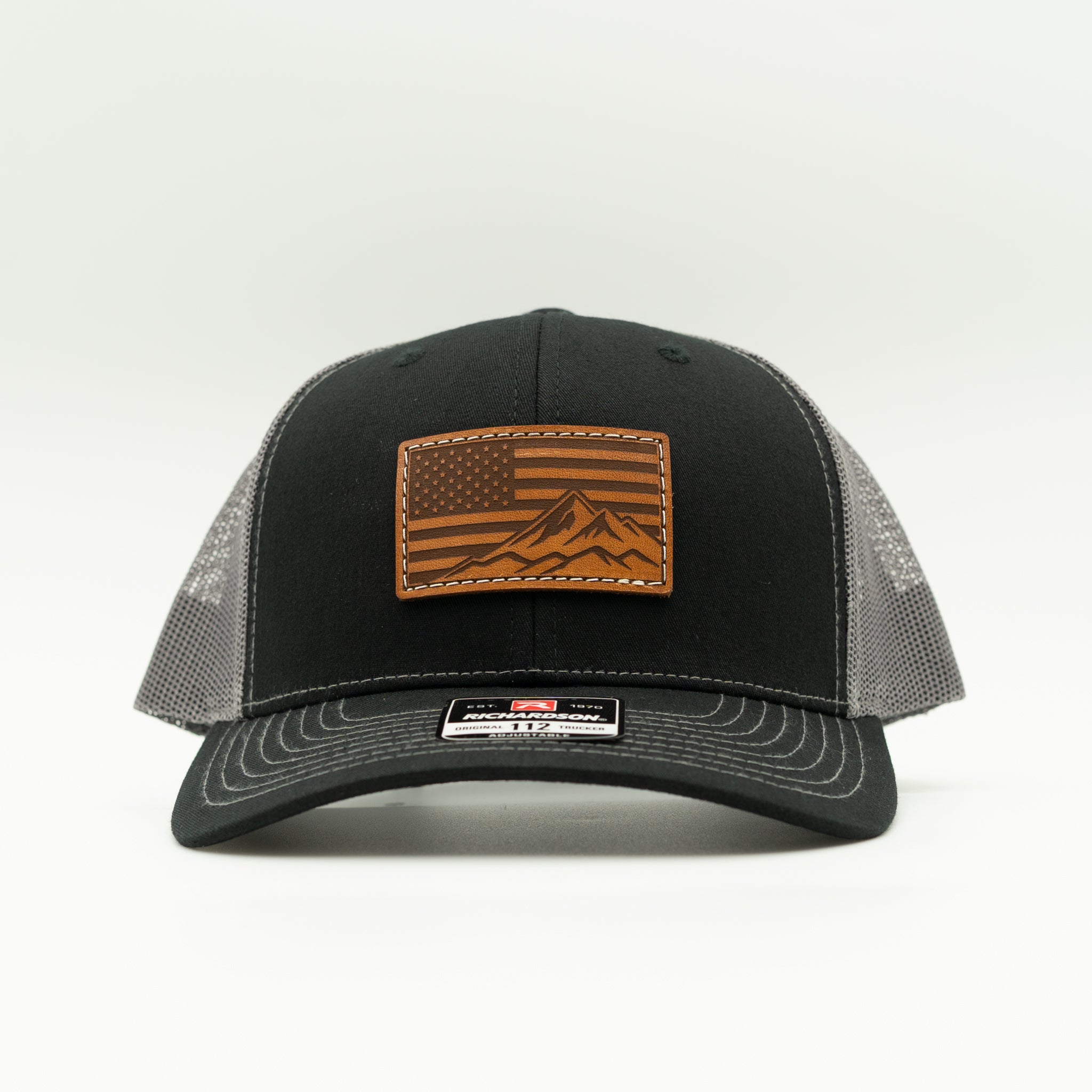 Front angled view of American Range leather patch trucker hat on black and charcoal Richardson 112, with light brown American flag mountain patch, curved bill, white stitching, mesh back, and snapback, a patriotic mountain hat for hiking and weekends.