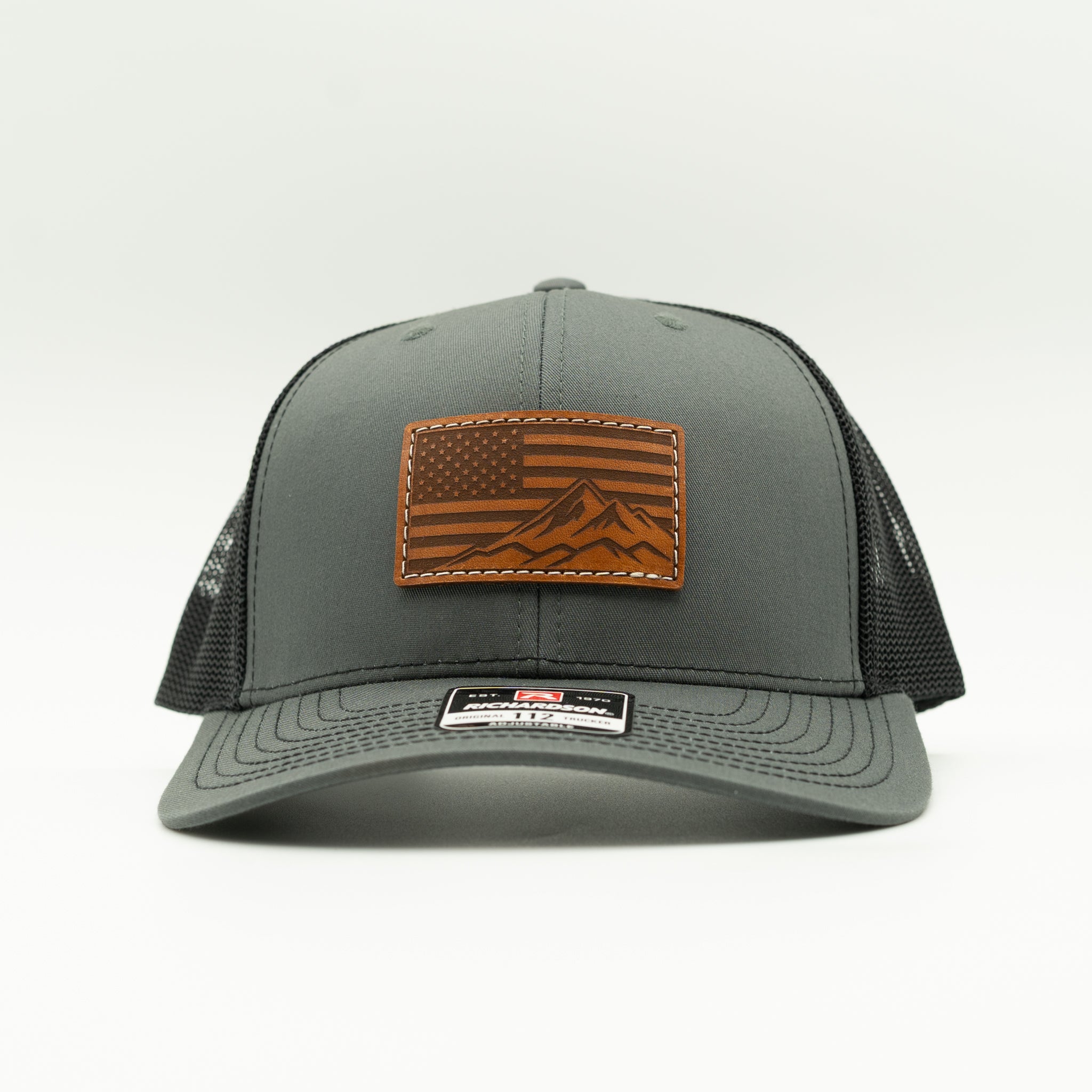 Front angled view of American Range leather patch trucker hat on charcoal and black Richardson 112, with light brown American flag mountain patch, curved bill, white stitching, mesh back, and snapback, a patriotic mountain hat for road trips and everyday wear.