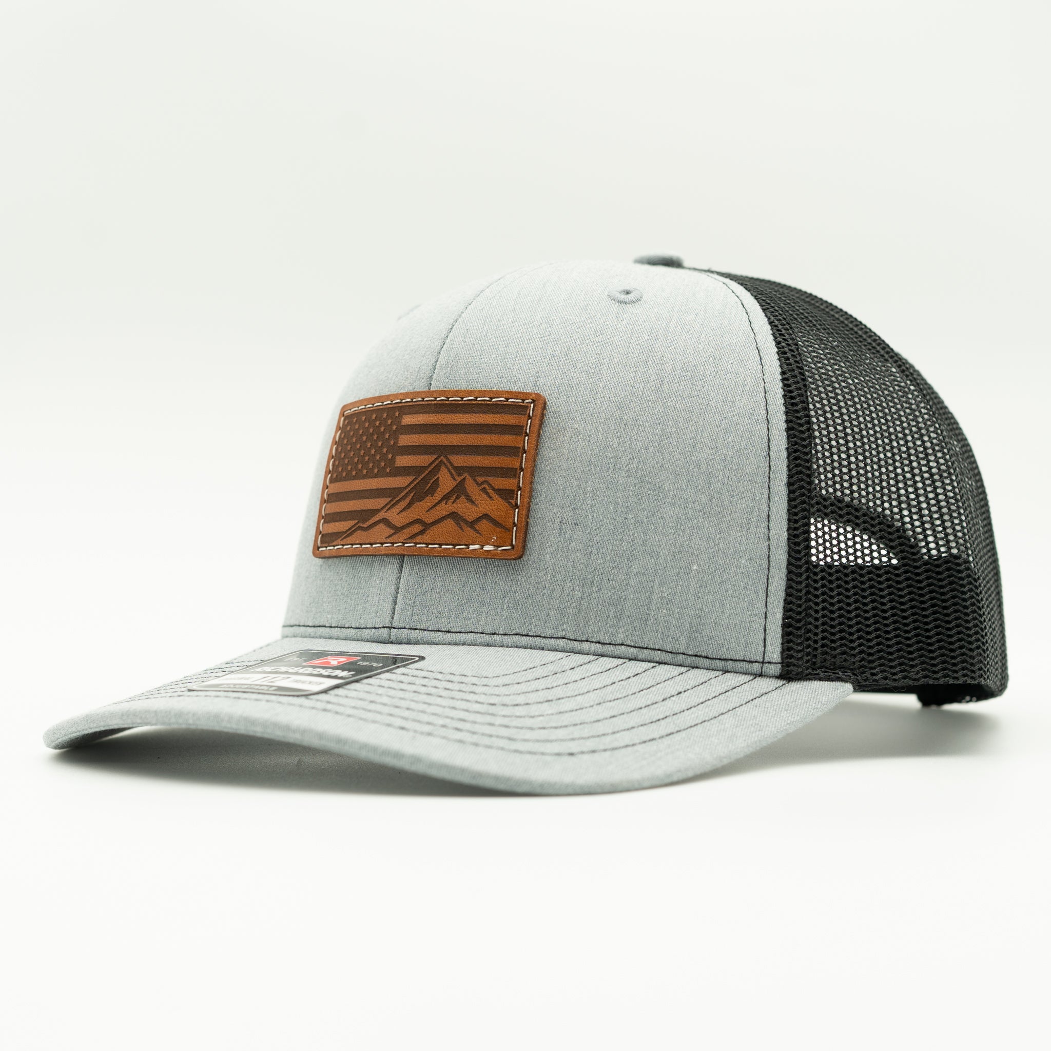 Side profile of American Range American flag leather patch hat on heather grey and black Richardson 112 trucker, showing patch, curved bill, mesh back, and snapback, a patriotic mountain hat that pairs with jeans, flannels, and outdoor gear.