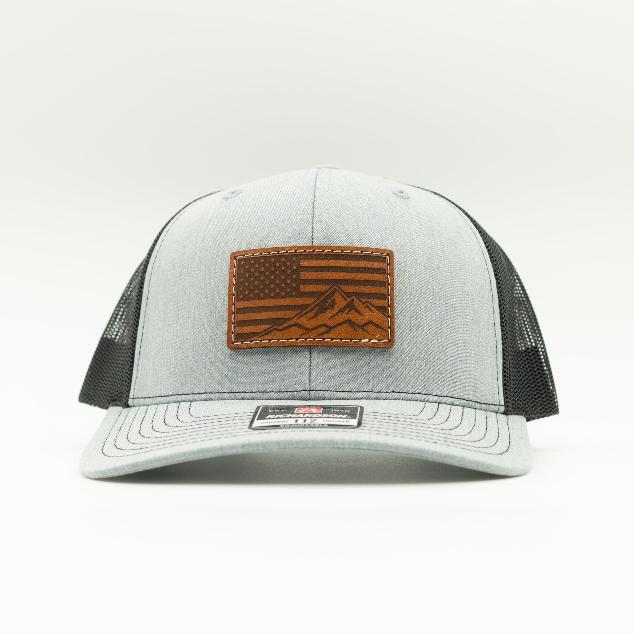 Front angled view of American Range leather patch trucker hat on heather grey and black Richardson 112, with light brown American flag mountain patch, curved bill, white stitching, mesh back, and snapback, a patriotic mountain hat for everyday casual wear.