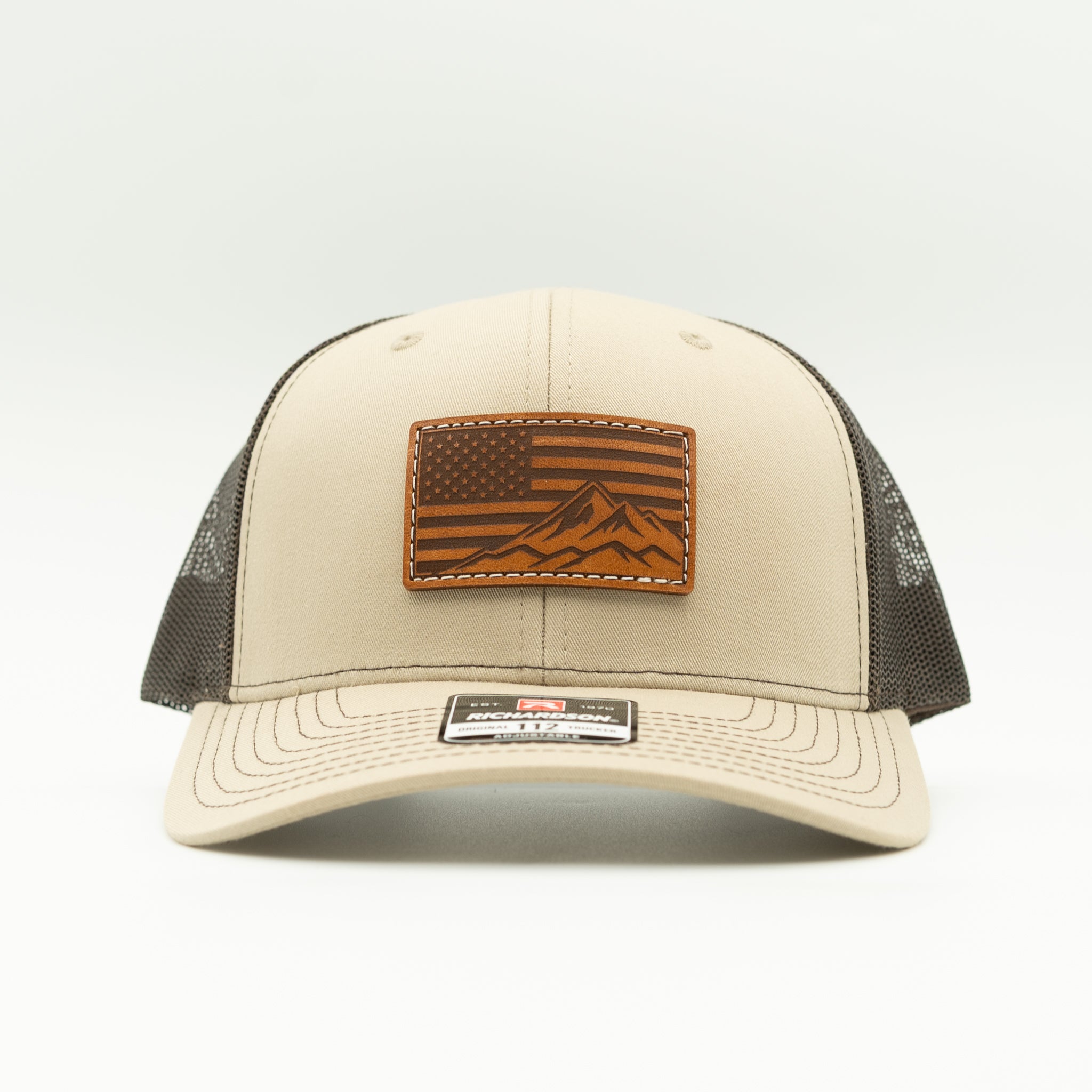 Front angled view of American Range leather patch trucker hat on khaki and brown Richardson 112, with light brown American flag mountain patch, curved bill, white stitching, mesh back, and snapback, a rugged patriotic mountain hat for camping and travel.