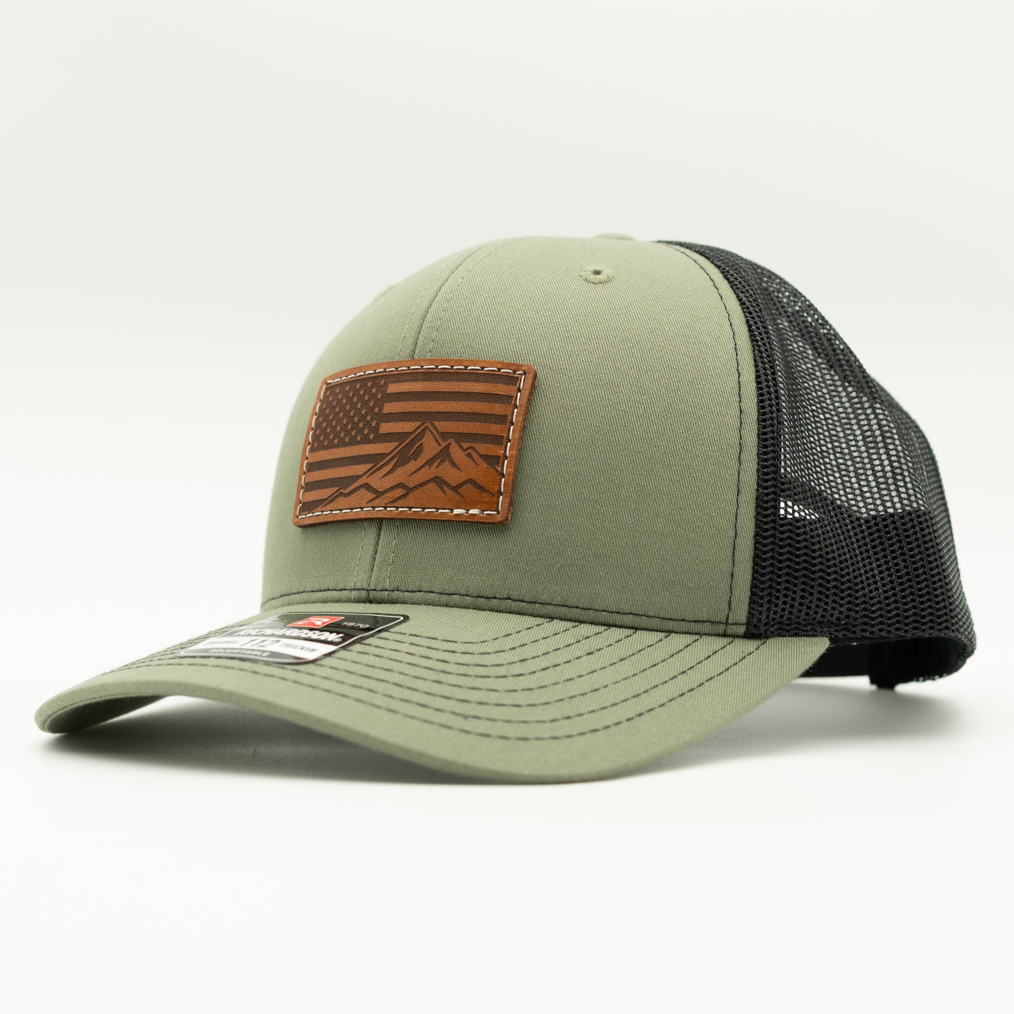 Side profile of American Range American flag leather patch hat on loden and black Richardson 112 trucker, showing light brown patch, curved bill, mid profile fit, mesh back, and snapback, a patriotic mountain hat for outdoor adventures.