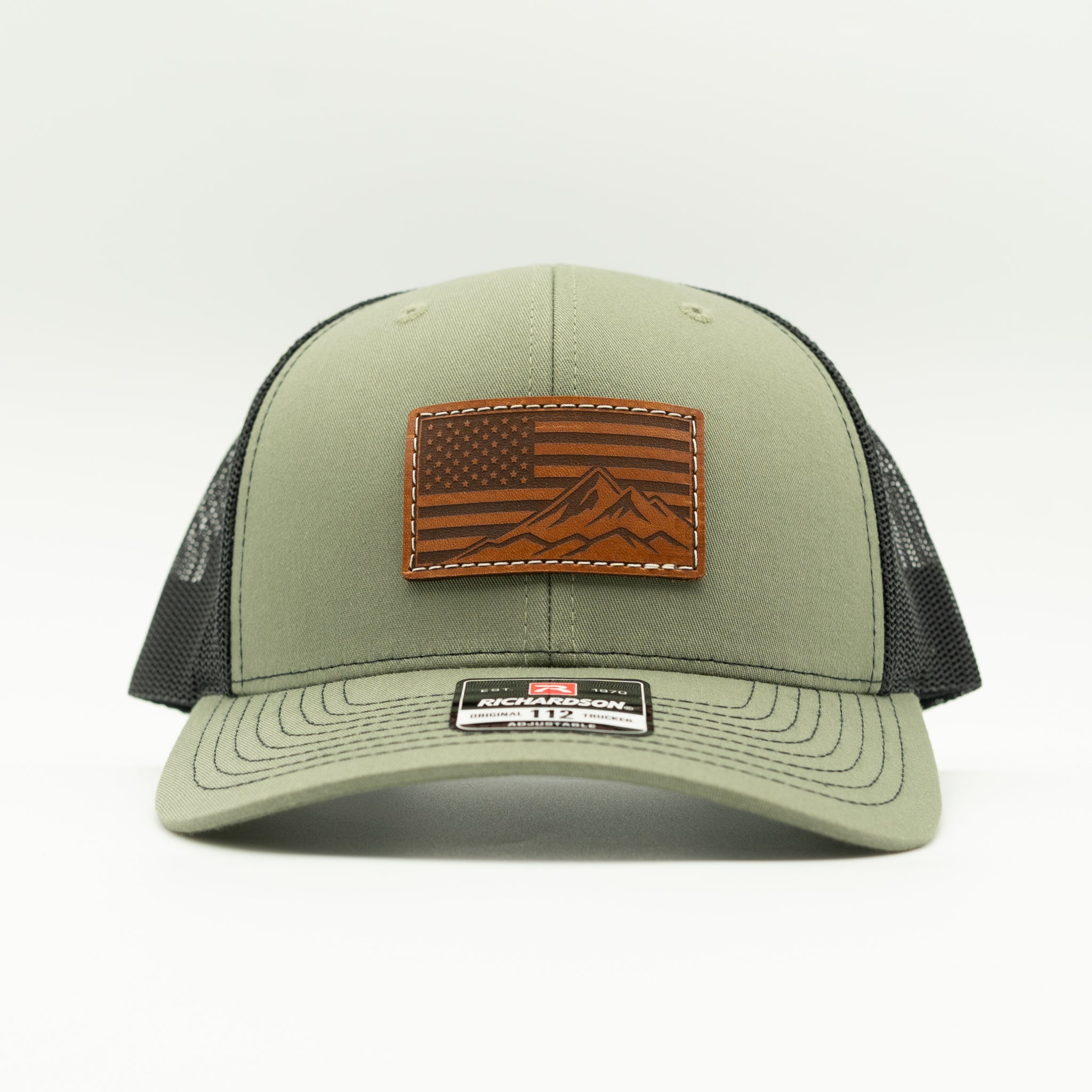 Front angled view of American Range leather patch trucker hat on loden and black Richardson 112, with light brown American flag patch, curved bill, white stitching, mesh back, and snapback, a patriotic mountain hat for hiking and everyday wear.