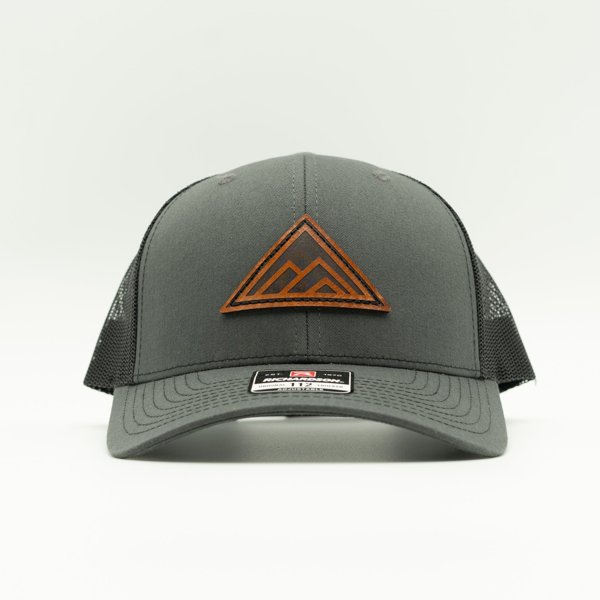 A dark gray trucker hat with black mesh back panels and a brown leather patch mountain logo on the front, shown against a plain white background.
Color: Charcoal/Black
