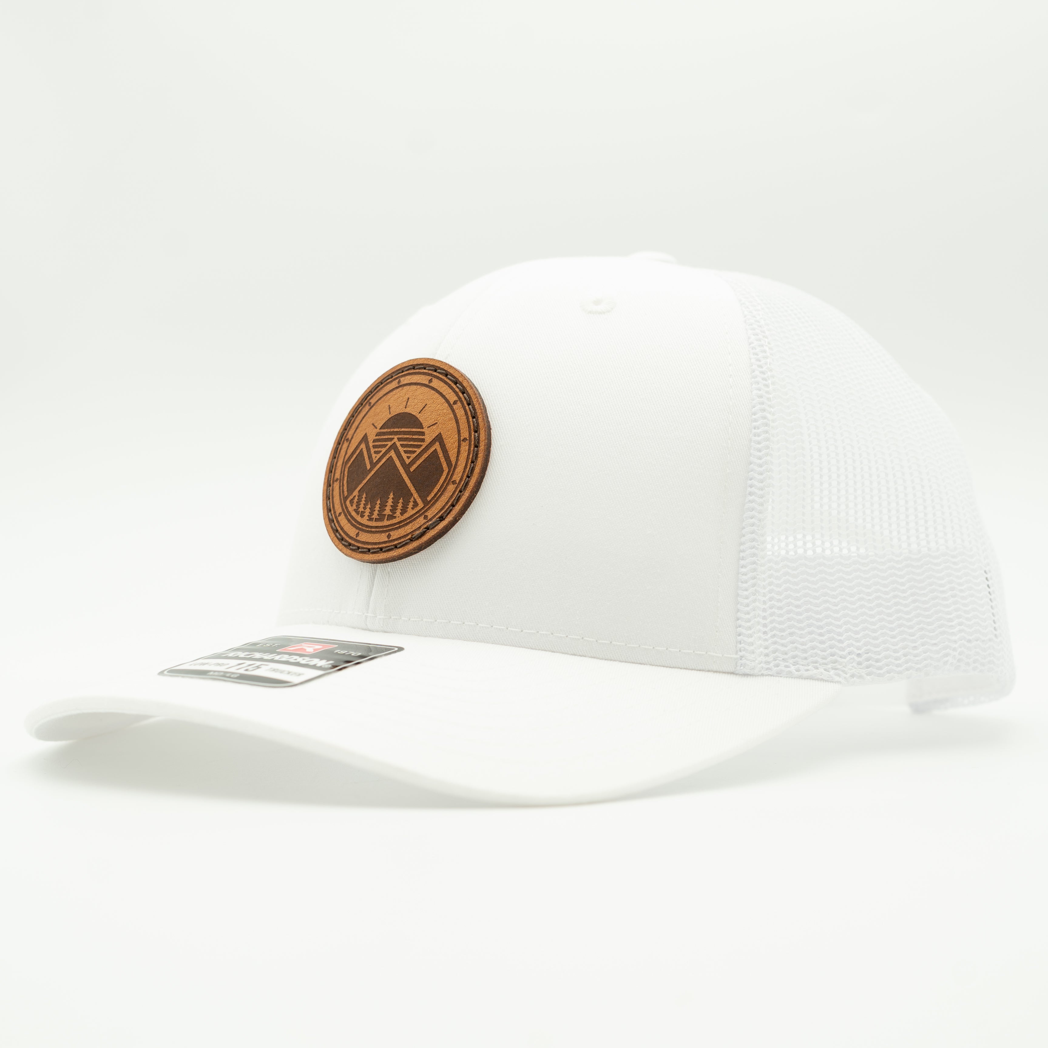 A white trucker-style baseball cap with a curved brim, mesh back panels, and a circular brown leather patch on the front featuring a mountain and tree design. A small retail sticker is visible on the brim.
Color: Solid White