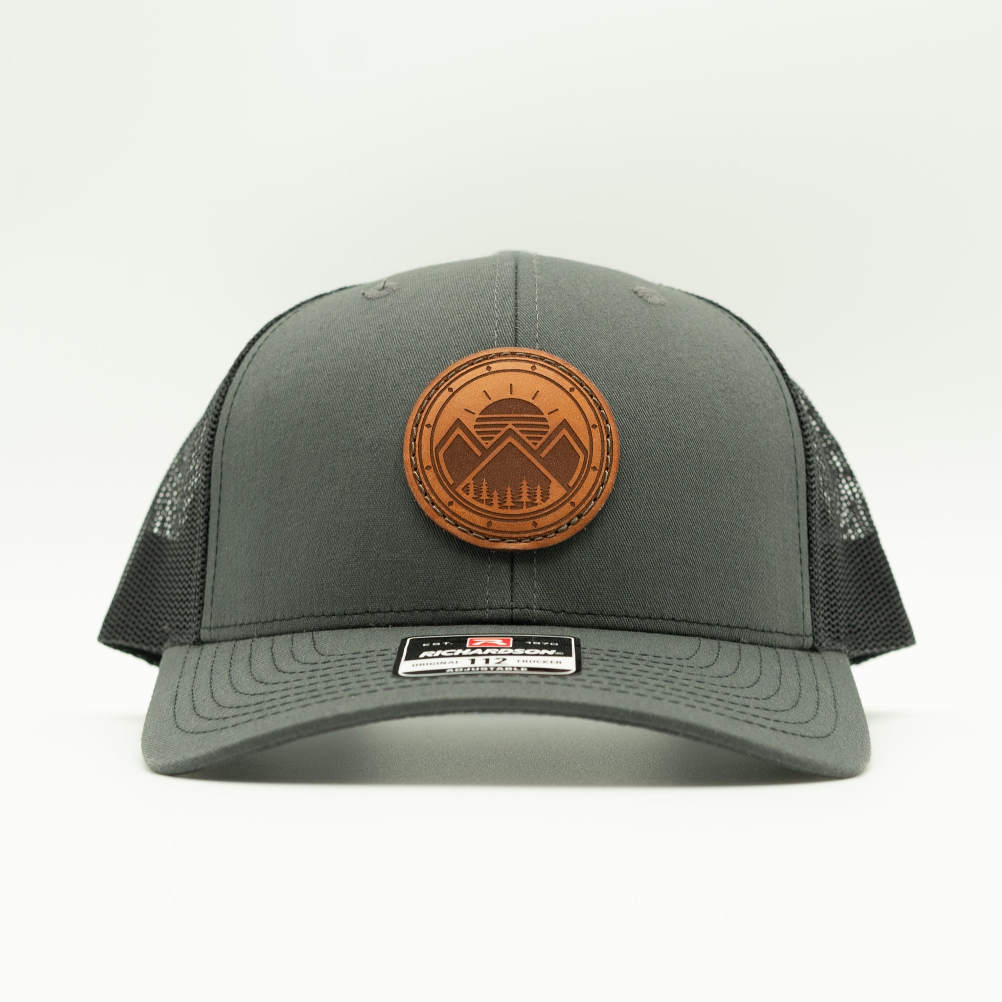 A dark gray trucker hat with a brown circular leather patch on the front featuring a mountain logo. The hat has a curved brim, dark gray front panel, and black mesh back panels, photographed against a clean white background.
Color: Charcoal/Black