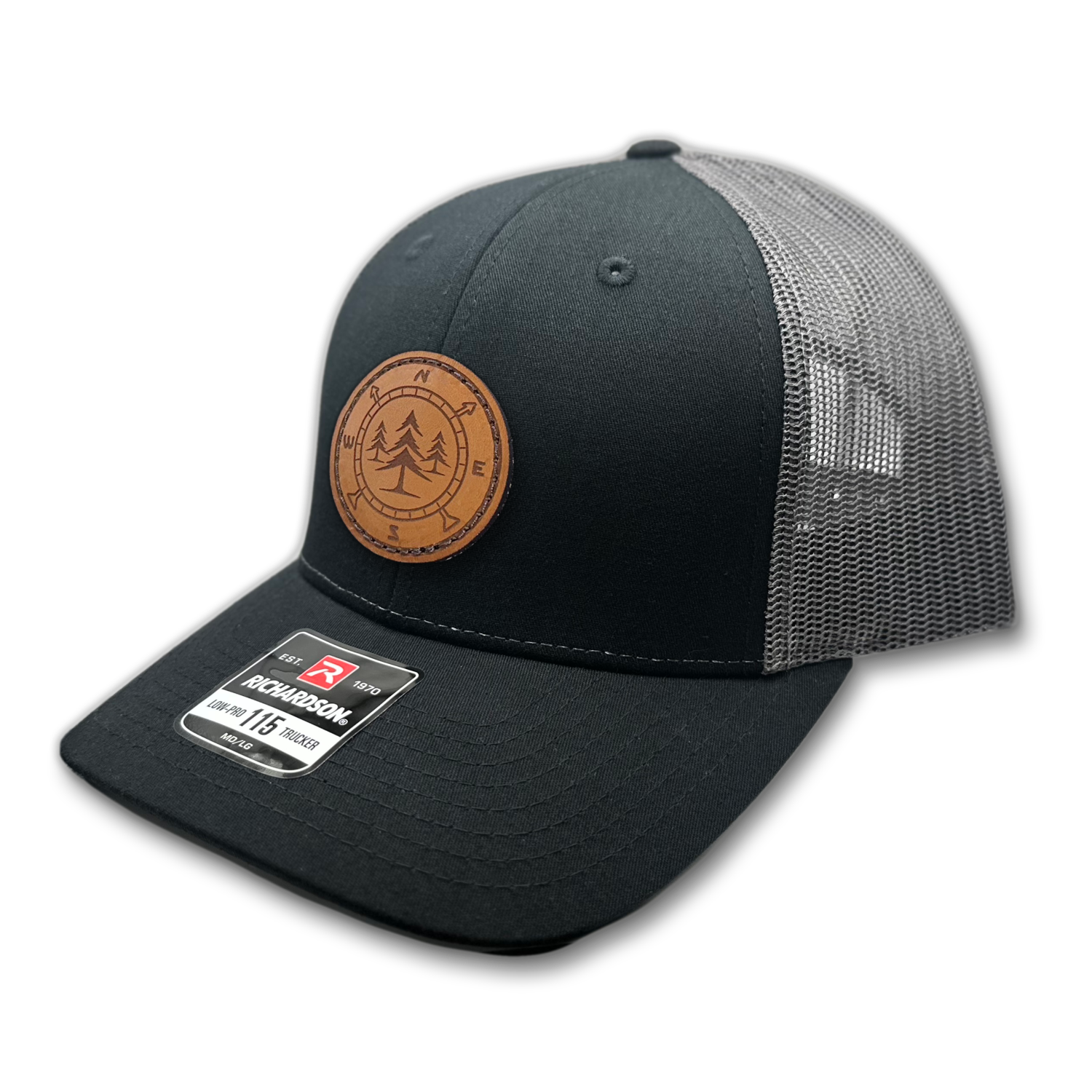 A close-up photo of a leather patch hat called Lost Pines. The hat features a unique compass and pine trees design on a circular leather patch that is sewn onto the front. The hat is a charcoal/black Richardson 115 low profile SnapBack adjustable hat, made in the USA. Available m/l sizes and multiple colors. Inspired by the mountains of Colorado, USA. 