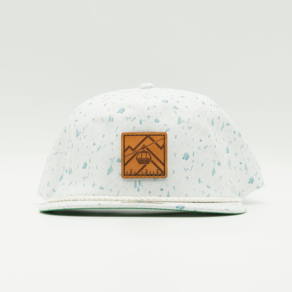 A white flat-brim cap with light blue speckles, featuring a brown leather patch on the front with an engraved mountain and cable car design.
Color: Blue Mountain