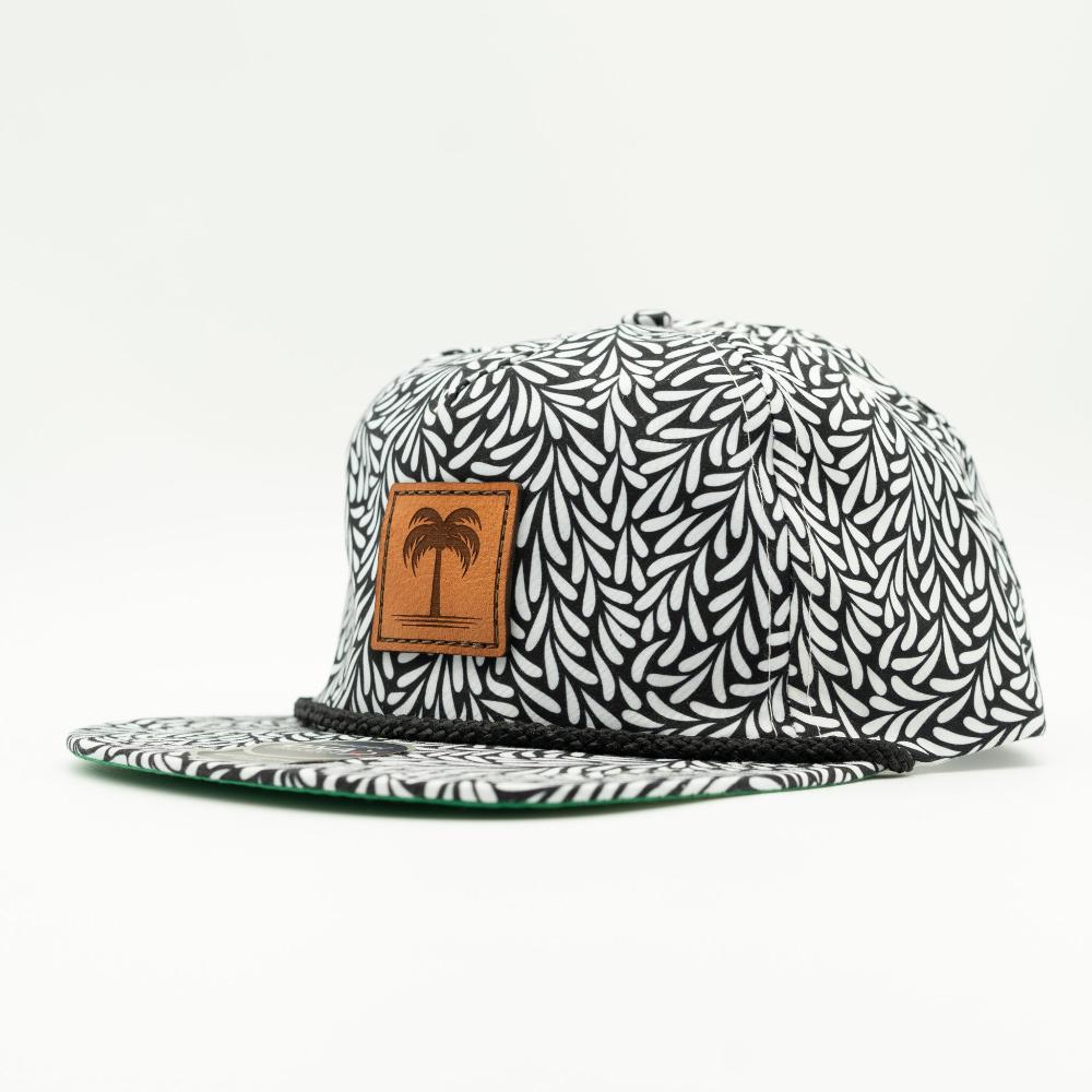 A flat-brim snapback cap featuring a black and white leaf pattern, with a brown leather patch on the front displaying a palm tree logo.
Color: Throwback Black