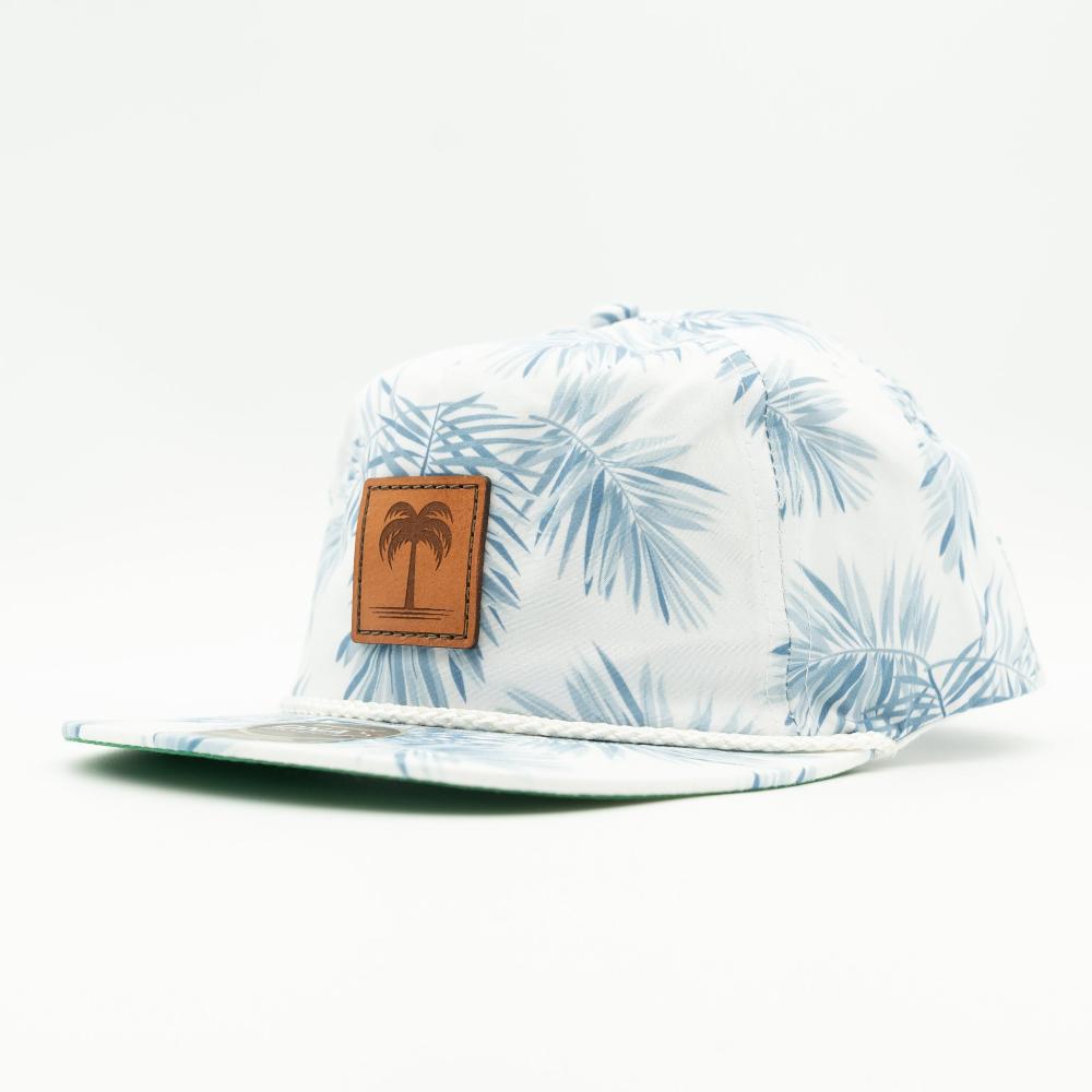 A white flat-brim cap with a light blue palm leaf pattern and a brown leather patch on the front featuring a palm tree logo, photographed on a clean white background.
Color: Floral Mist