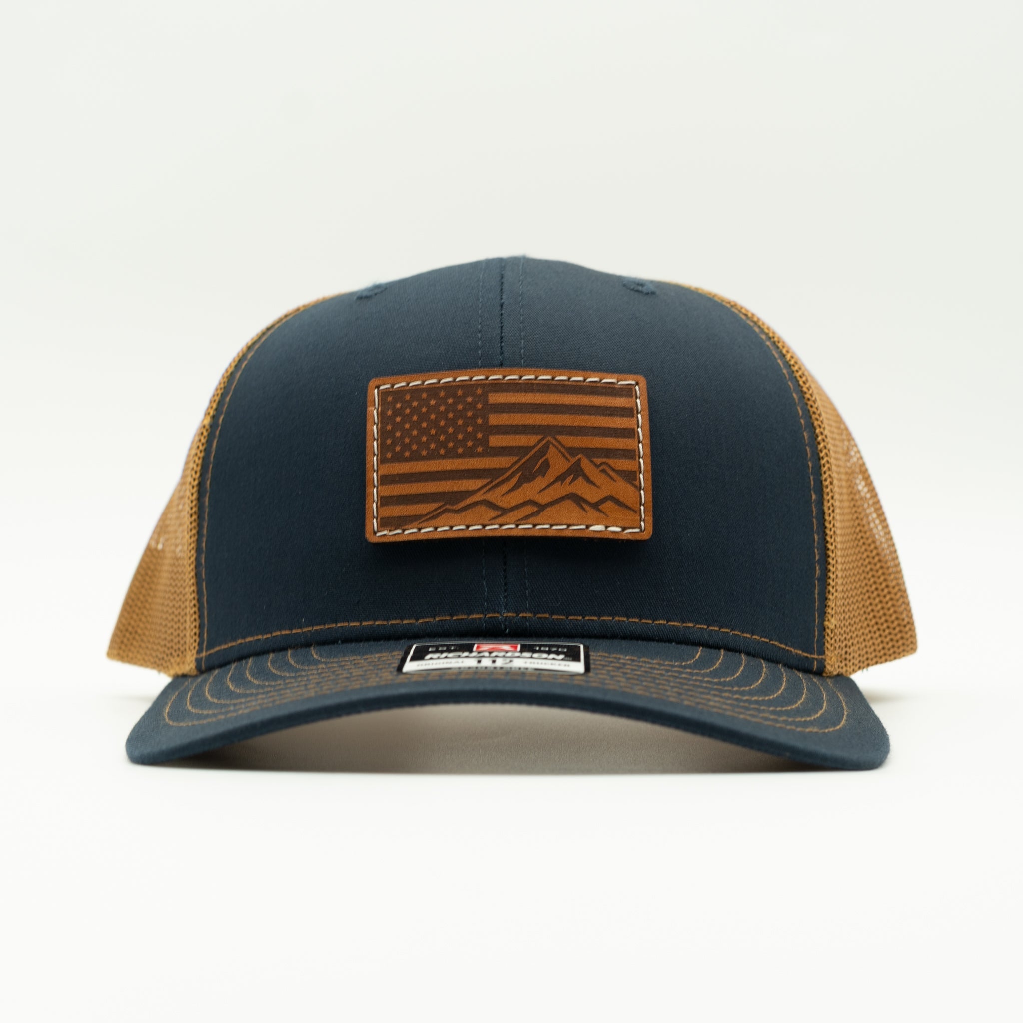 A navy blue and tan trucker cap with a brown leather patch featuring an American flag and mountain design on the front, set against a plain white background.
Color: Navy/Caramel