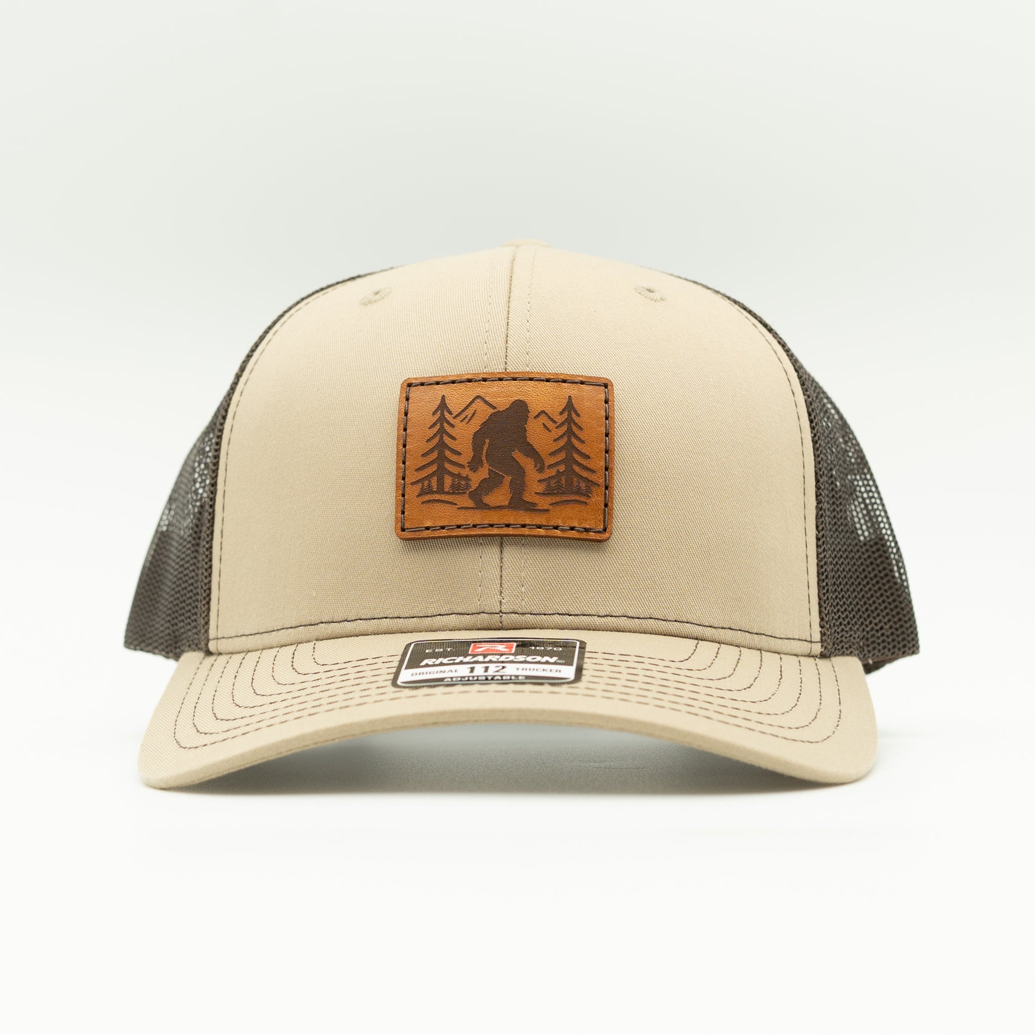 Front view of khaki/coffee Mountain Myth leather patch trucker hat on Richardson 112, Sasquatch and mountain design on veg-tan patch.