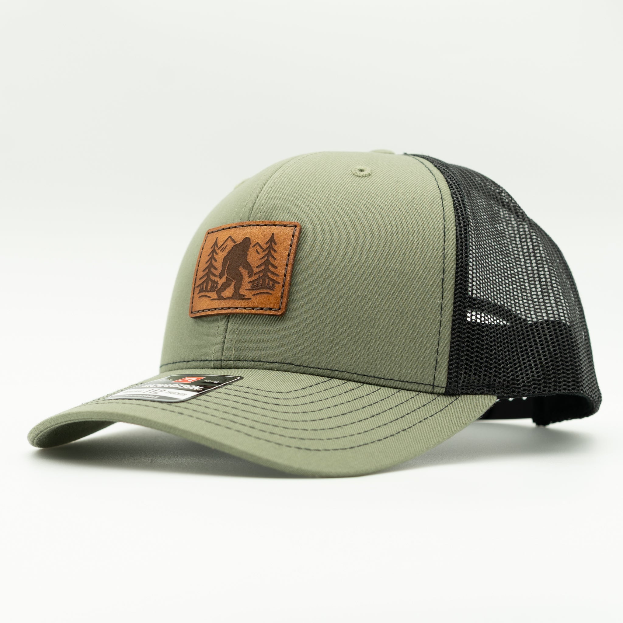 Side profile of loden green front, black mesh Richardson 112 Mountain Myth hat with brown Bigfoot leather patch and curved bill.