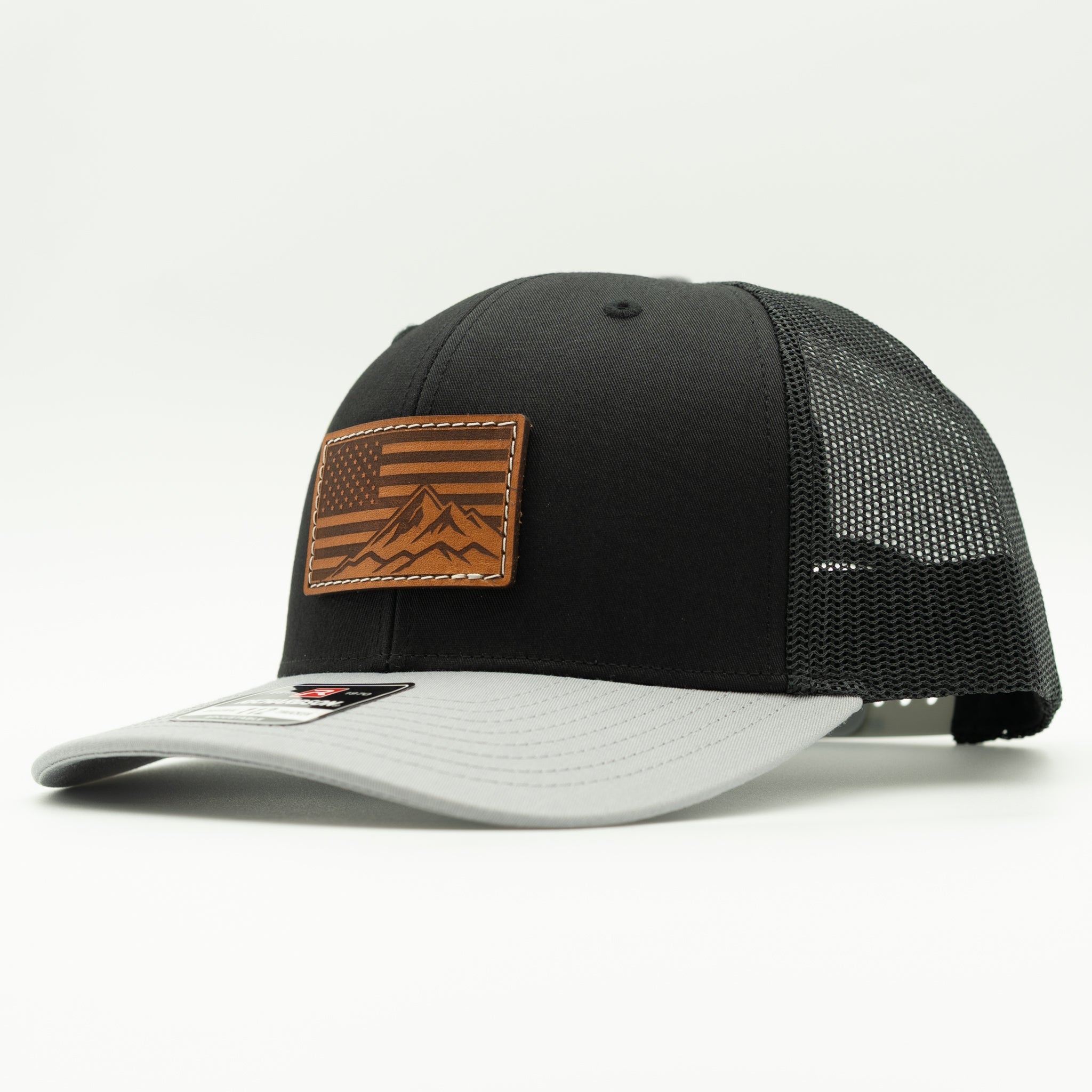 A black trucker-style cap with a light gray visor and black mesh back, featuring a brown leather patch on the front with an American flag and mountain design.
Color: Black/Grey Combo