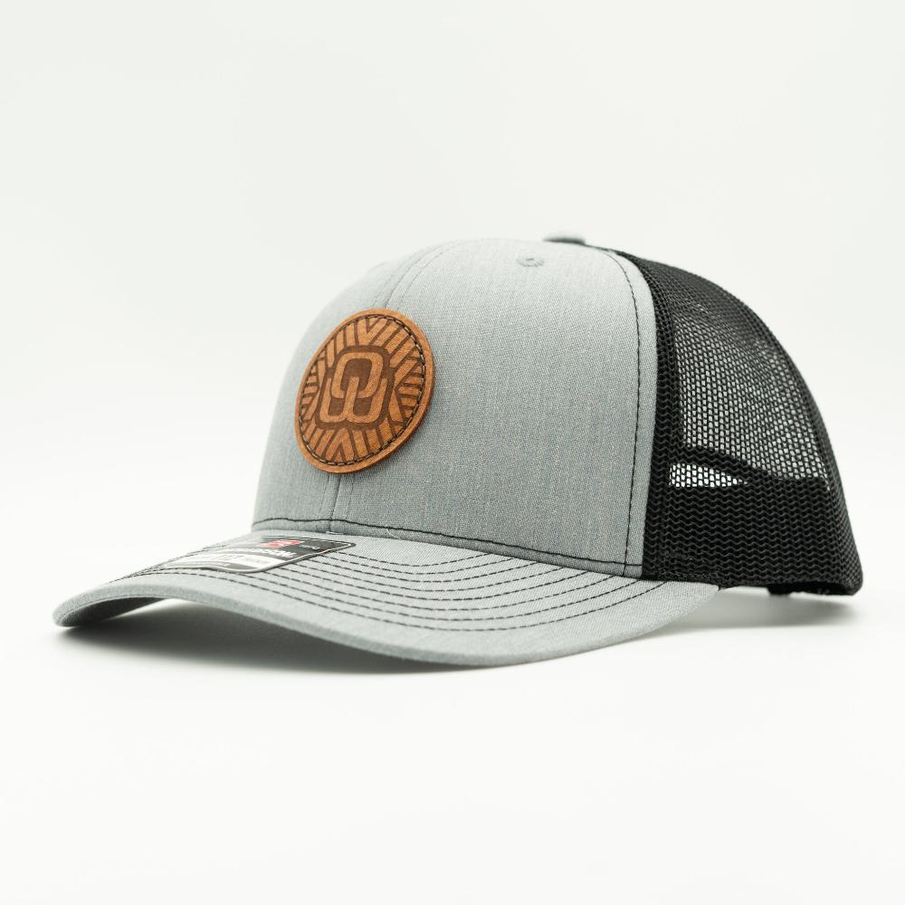 A light gray and black trucker cap with a brown circular leather patch on the front, photographed on a clean white background.
Color: Heather Grey/Black