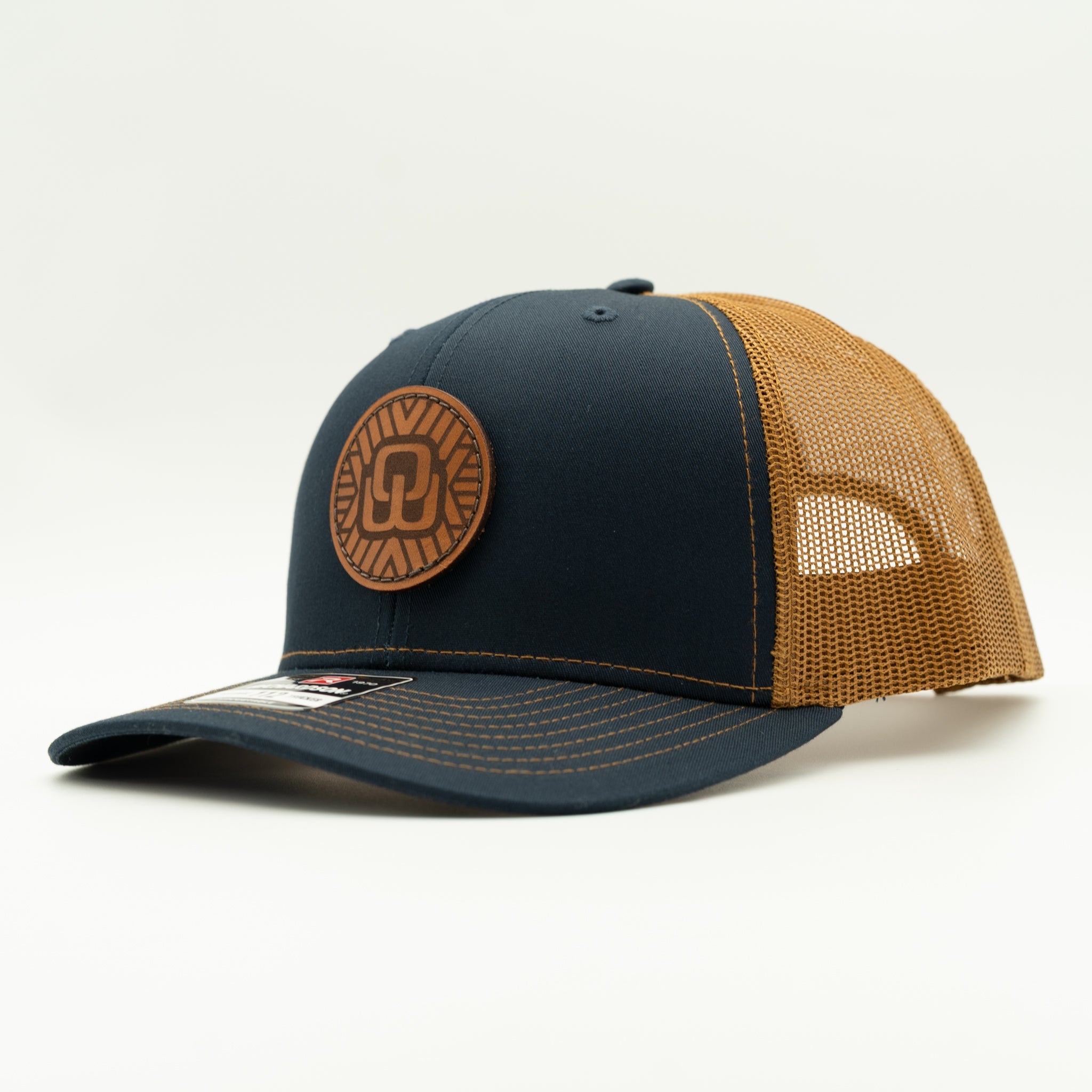 A trucker-style baseball cap with a dark navy blue front panel and brim, tan mesh back panels, and a circular brown leather patch on the front.
Color: Navy/Caramel