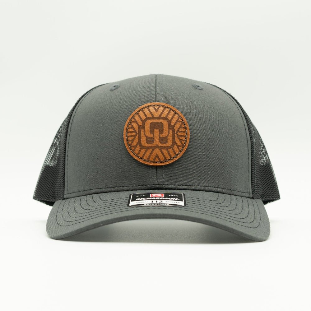 A dark gray trucker hat with a brown circular leather patch on the front and black mesh side panels, shown against a plain white background.
Color: Charcoal/Black