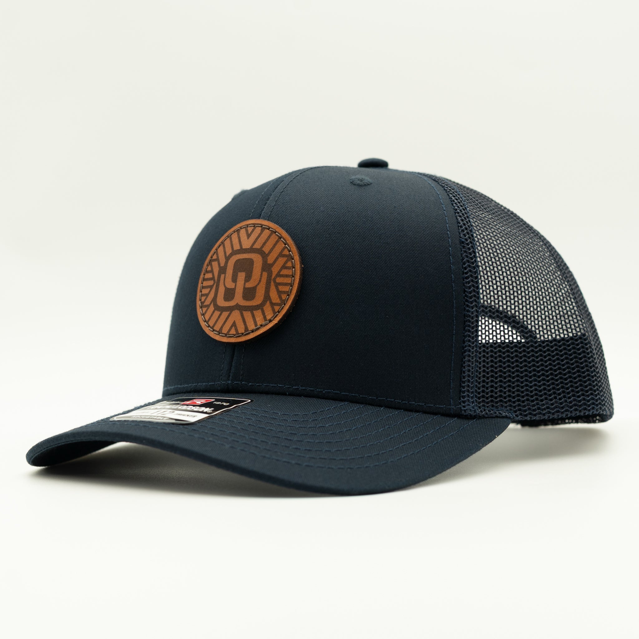 A dark navy blue trucker-style baseball cap with a brown circular leather patch on the front, set against a plain light background.
Color: Navy/Navy