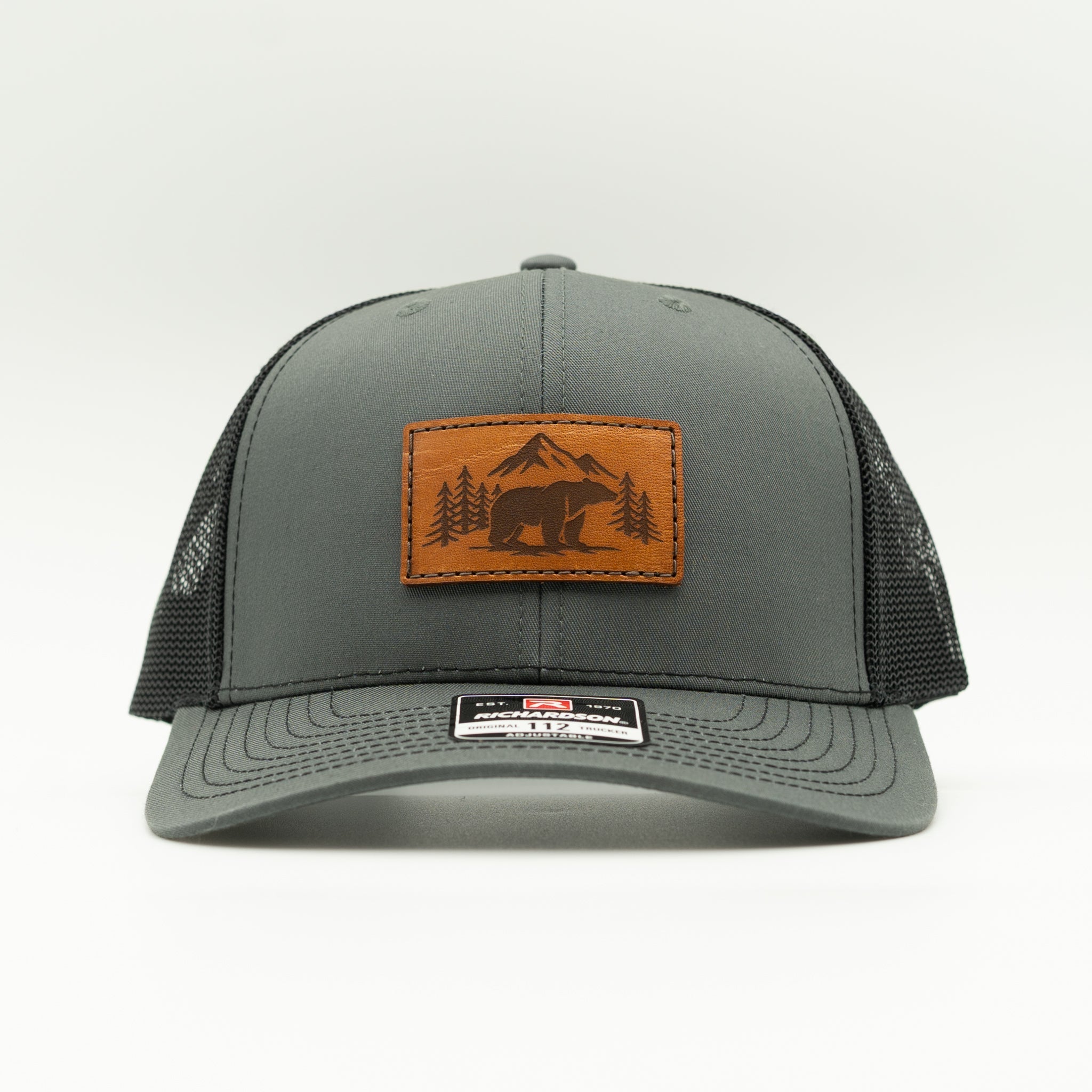 Front view of Bearline Peak leather patch hat on charcoal and black Richardson 112 trucker, showing engraved bear and pine tree leather patch, curved bill, and mesh back.
Color: Charcoal/Black