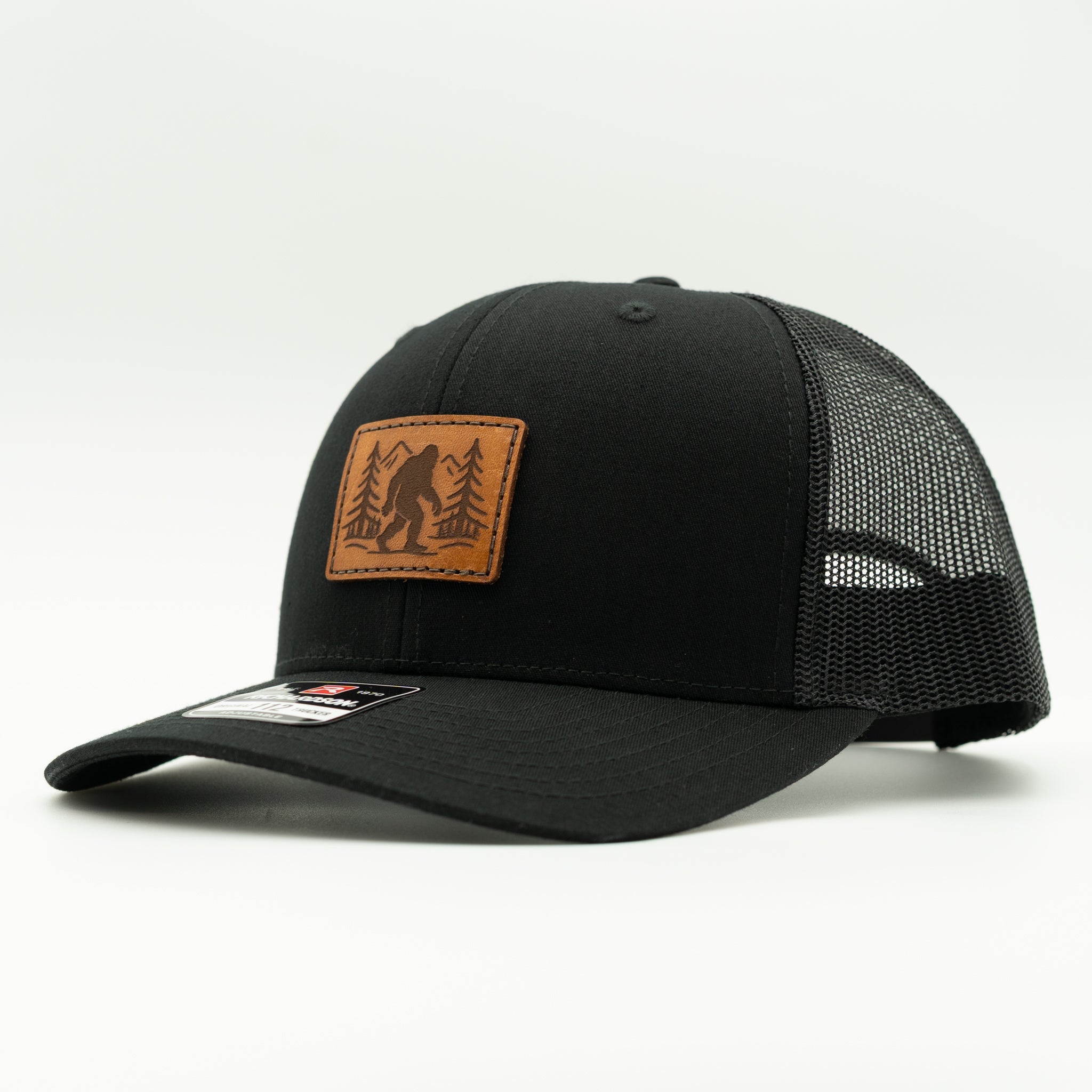 Side view of solid black Richardson 112 Mountain Myth leather patch hat with Bigfoot forest design, mesh back, and pre-curved visor.