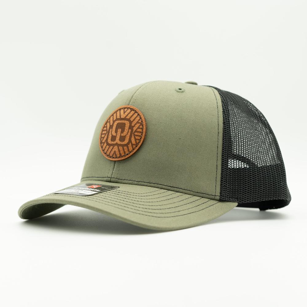 An olive green and black trucker cap with a circular brown leather patch on the front, photographed on a clean white background.
Color: Loden/Black