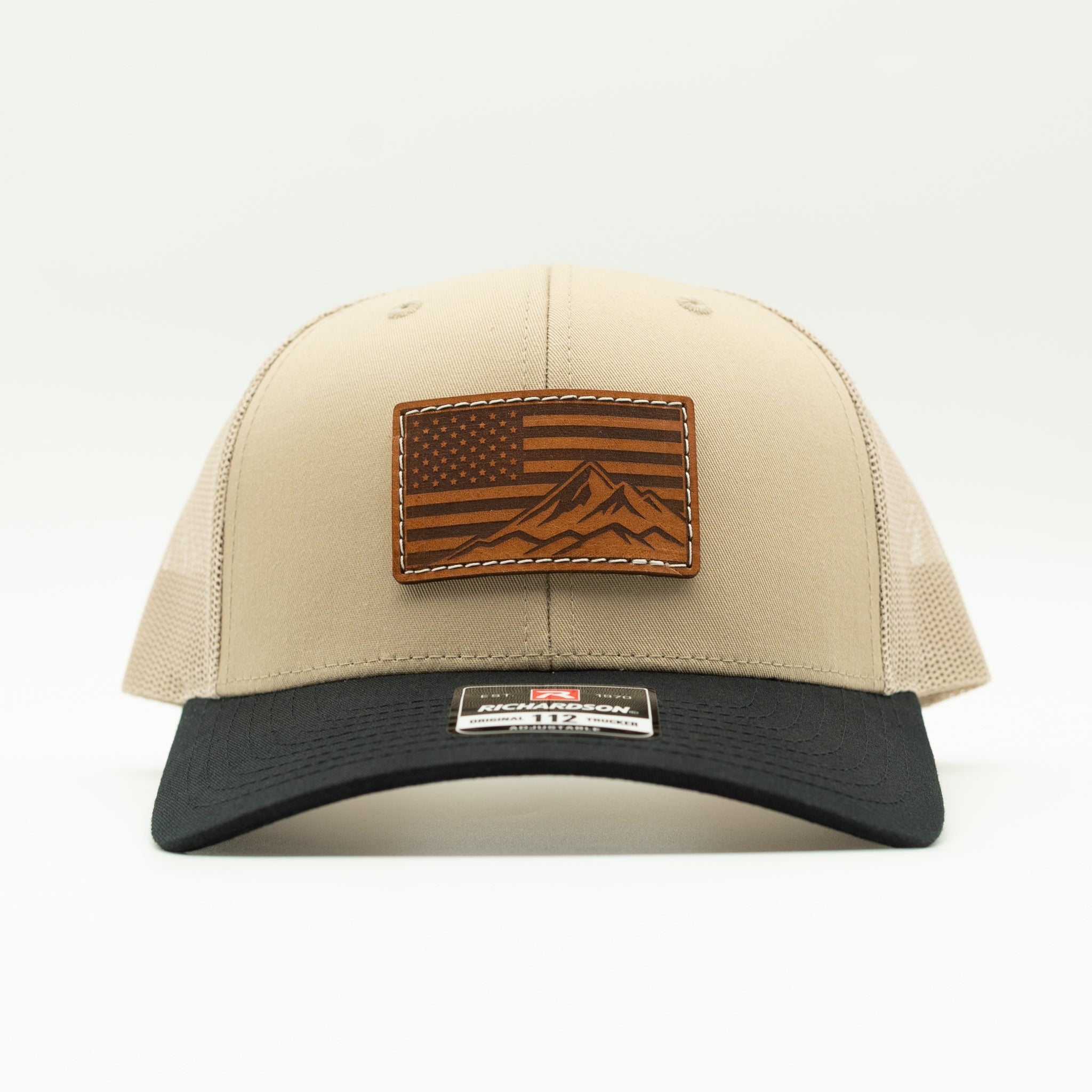 A beige and black trucker cap with a brown leather patch on the front featuring an American flag and mountain design, shown against a plain white background.
Color: Khaki/Black Combo
