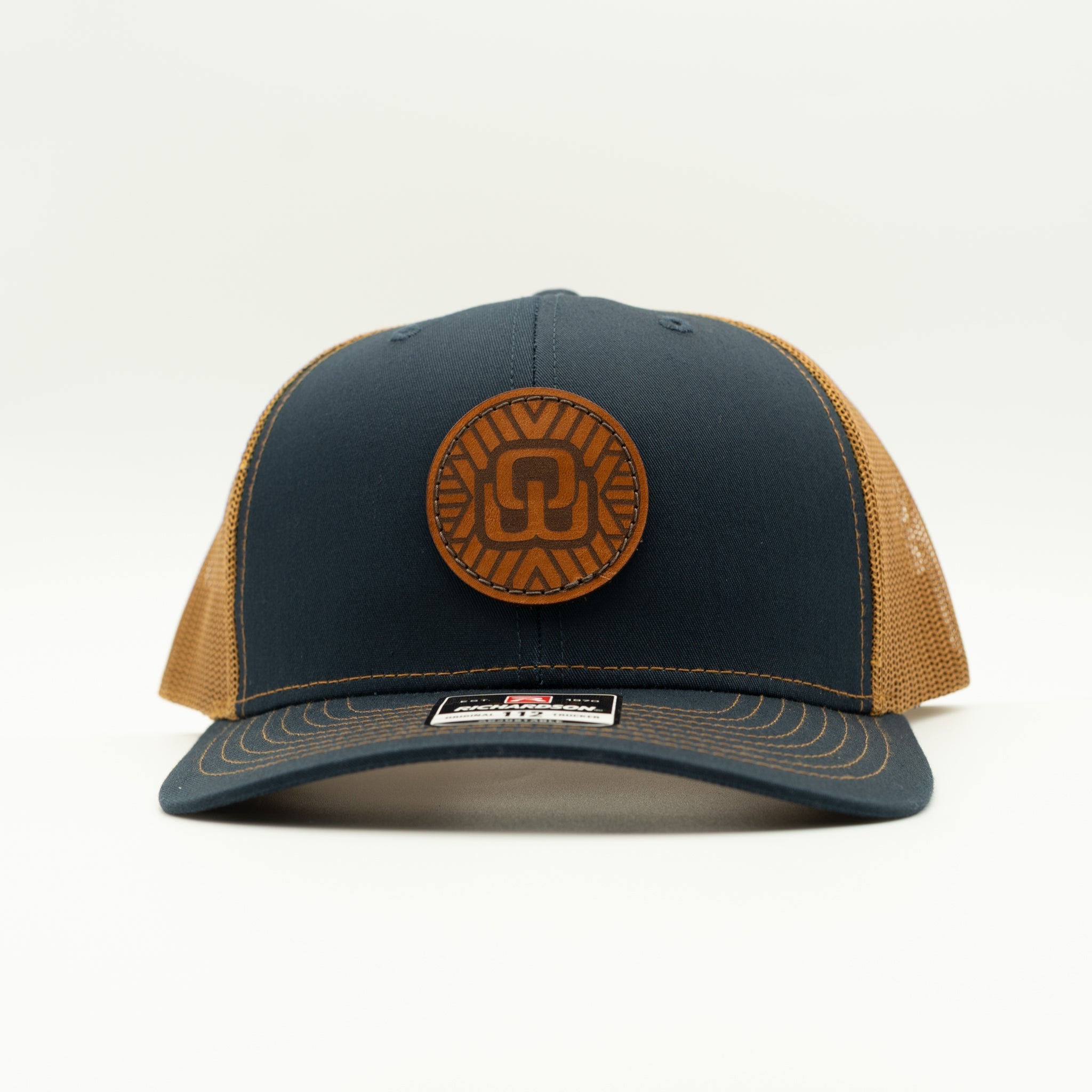 A dark navy blue trucker hat with tan mesh back panels and a circular tan leather patch on the front.
Color: Navy/Caramel