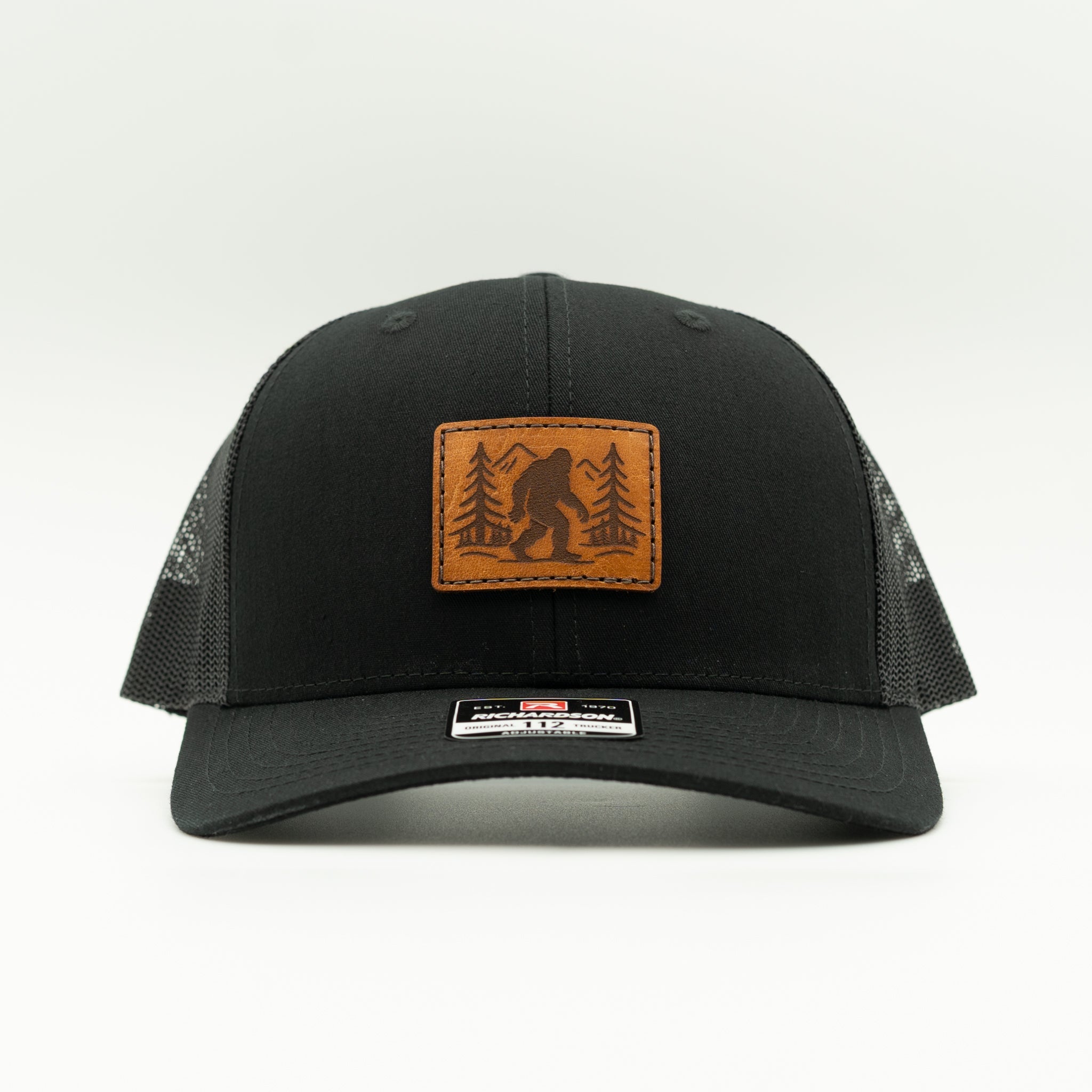 Front view of solid black Mountain Myth Bigfoot leather patch trucker hat on Richardson 112, structured mid-profile crown and curved bill.