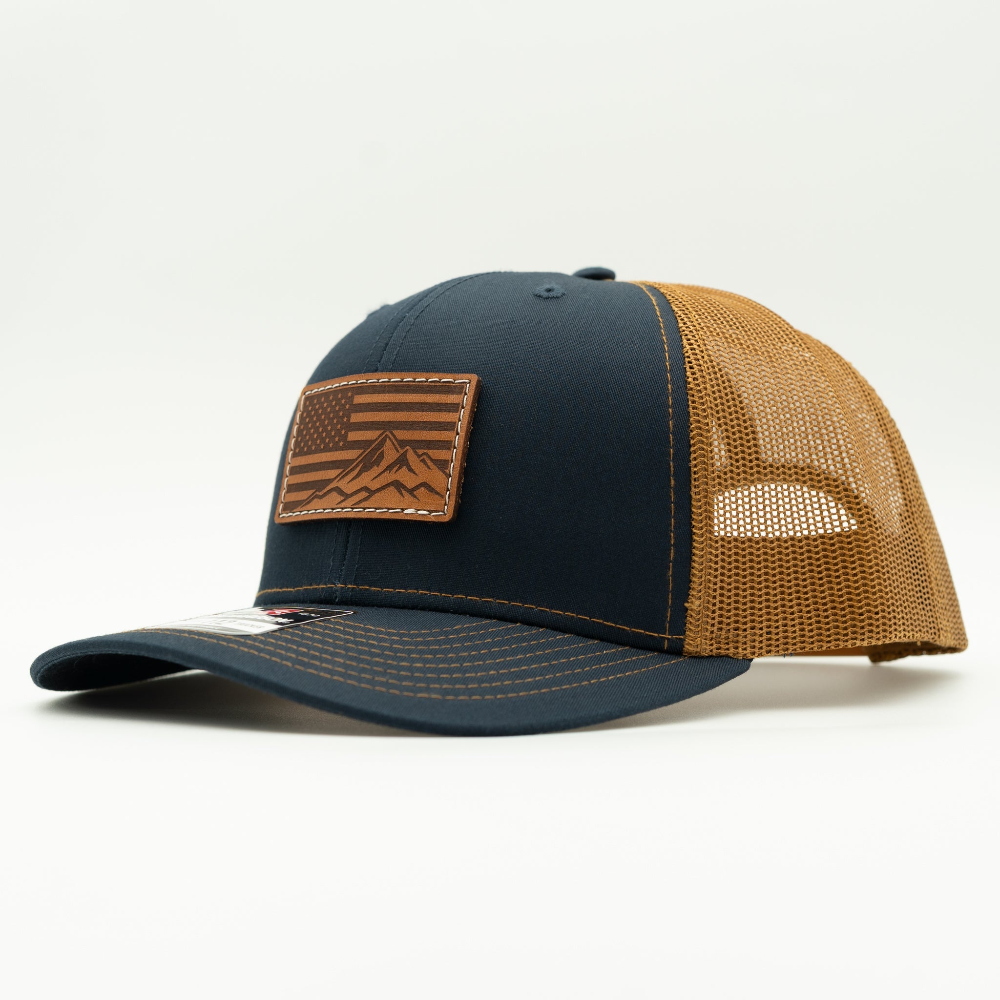 A structured trucker-style baseball cap with a dark navy front panel and brim, tan mesh back panels, and a brown leather patch on the front featuring a mountain and flag design.
Color: Navy/Caramel