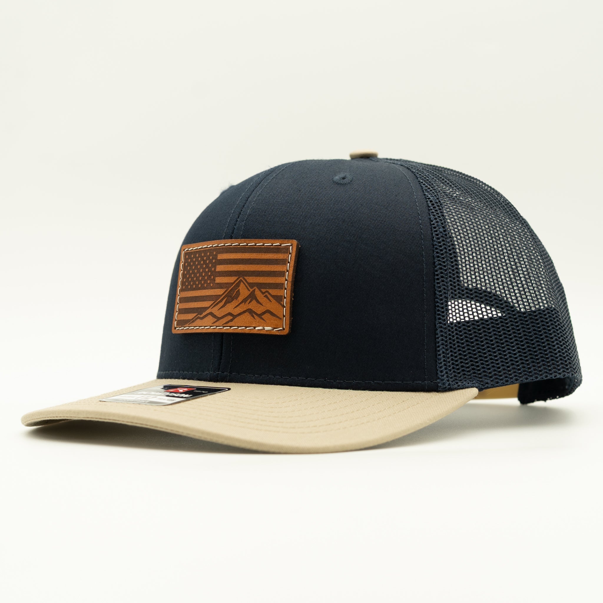 A trucker-style baseball cap with a dark navy blue front panel and mesh back, a beige flat brim, and a brown leather patch on the front featuring an American flag and mountain design.
Color: Navy/Khaki Combo