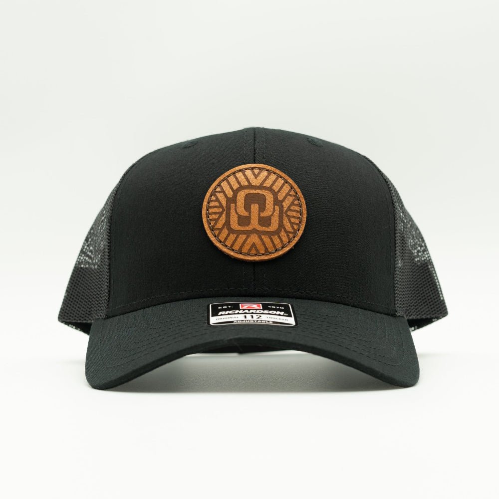 A black trucker hat with a brown circular leather patch on the front and black mesh side panels, shown against a plain white background.
Color: Solid Black