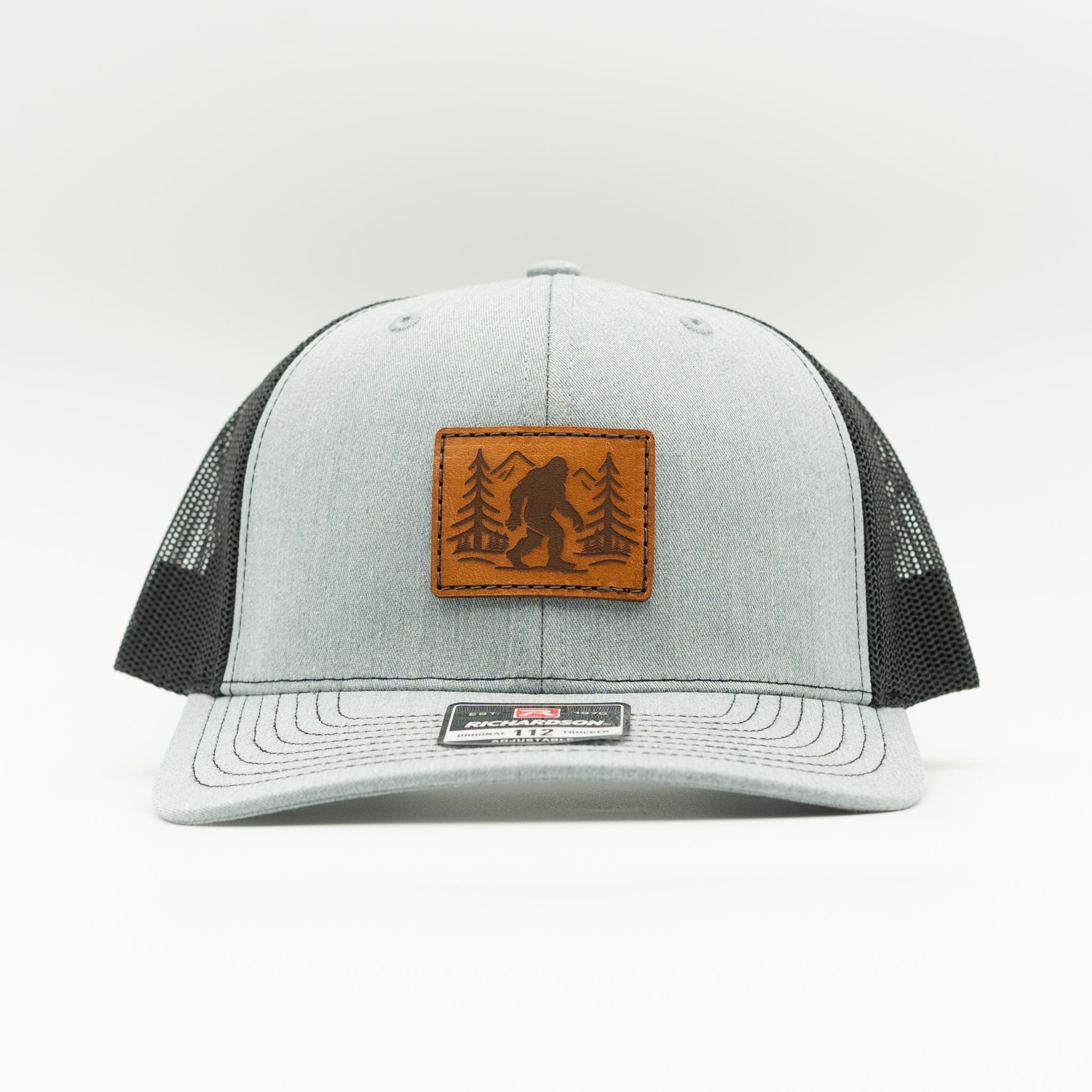 Front view of heather/black Mountain Myth leather patch trucker hat, Bigfoot forest design centered on structured Richardson 112 crown.