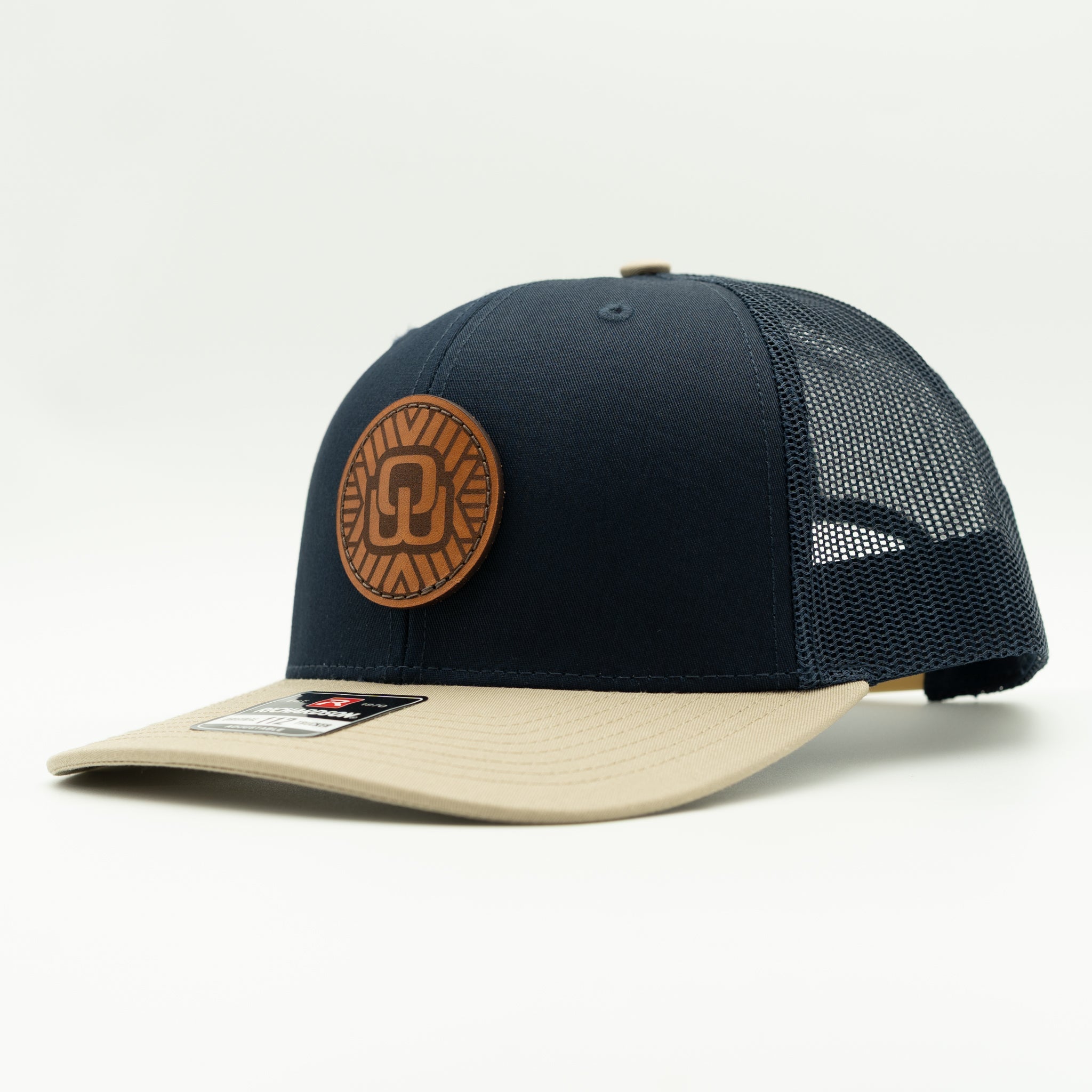 A navy blue and beige trucker hat with a circular brown leather patch on the front, photographed on a clean white background.
Color: Navy/Khaki Combo