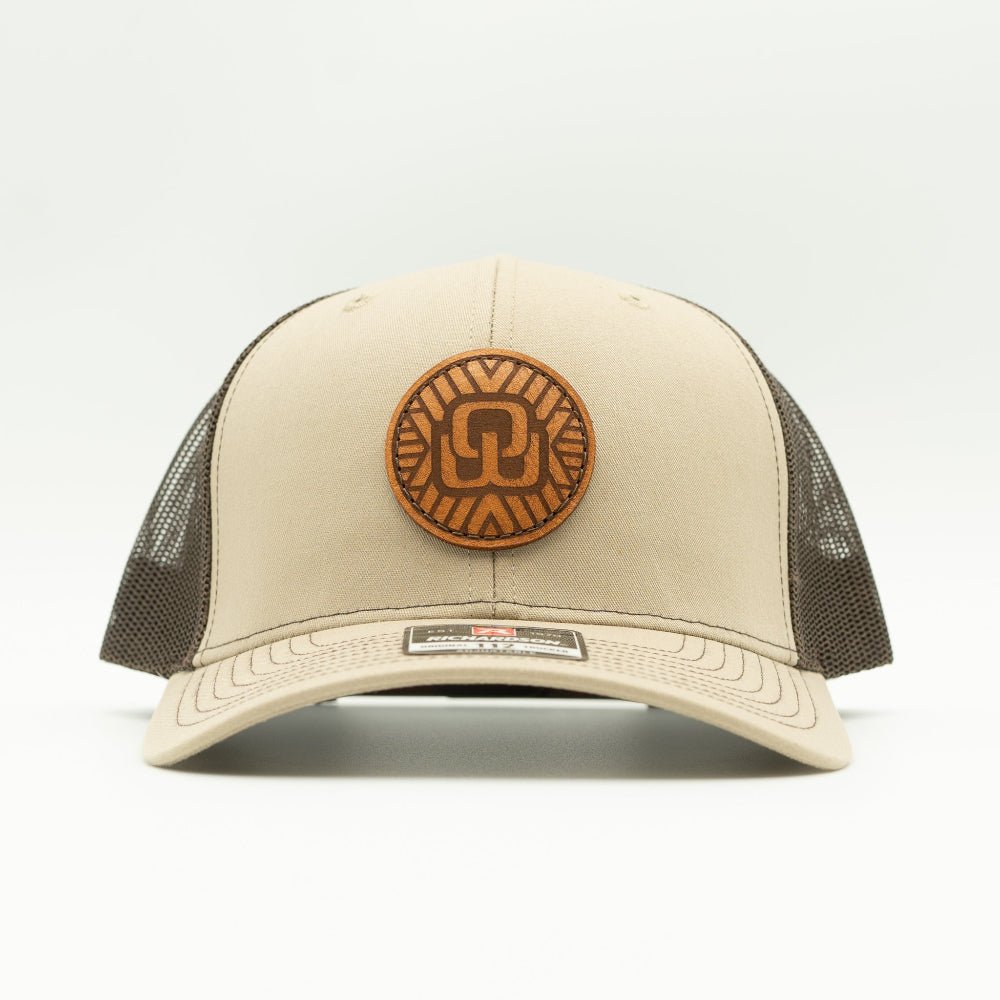 A beige trucker hat with dark brown mesh side panels and a circular brown leather patch on the front.. The hat is photographed against a clean, light gray background.
Color: Khaki/Coffee