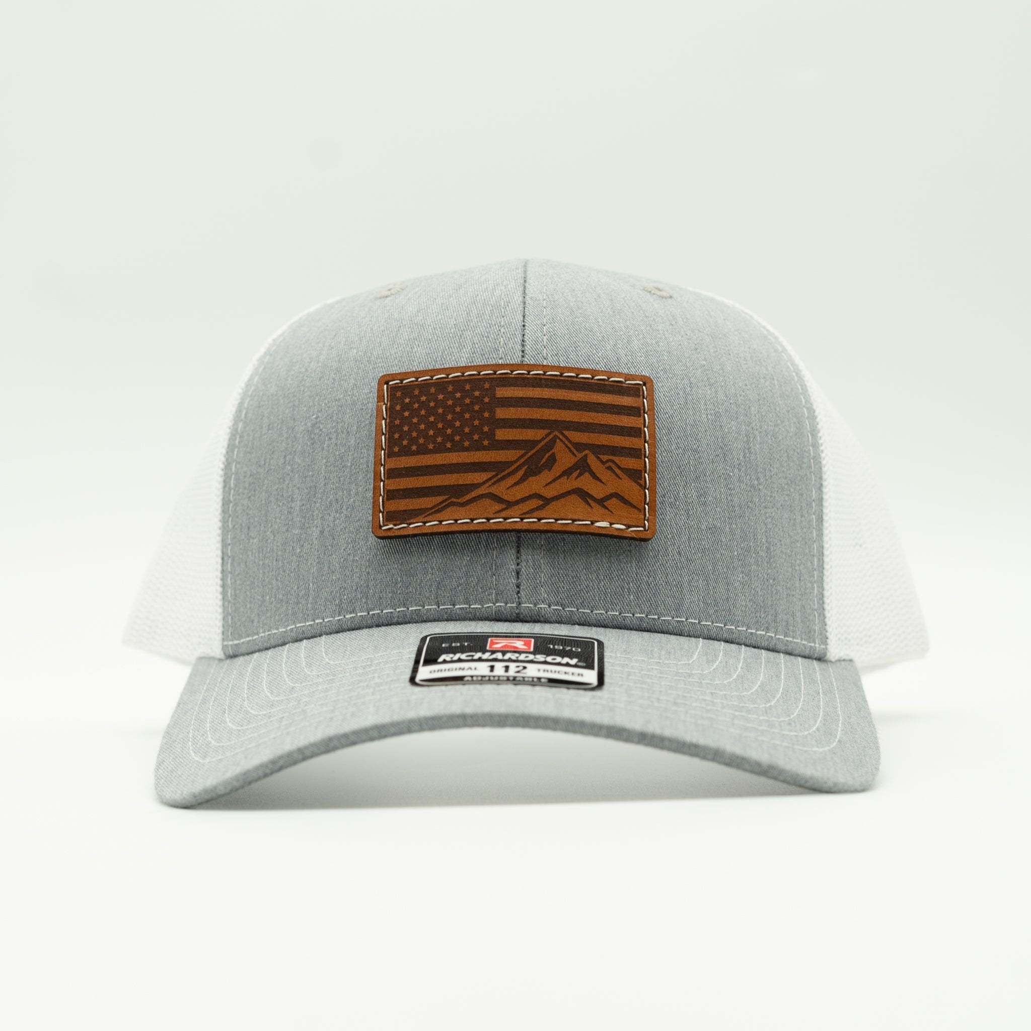 A light gray and white trucker cap featuring a brown leather patch on the front with an American flag and mountain design, photographed against a clean white background.
Color: Heather Grey/White